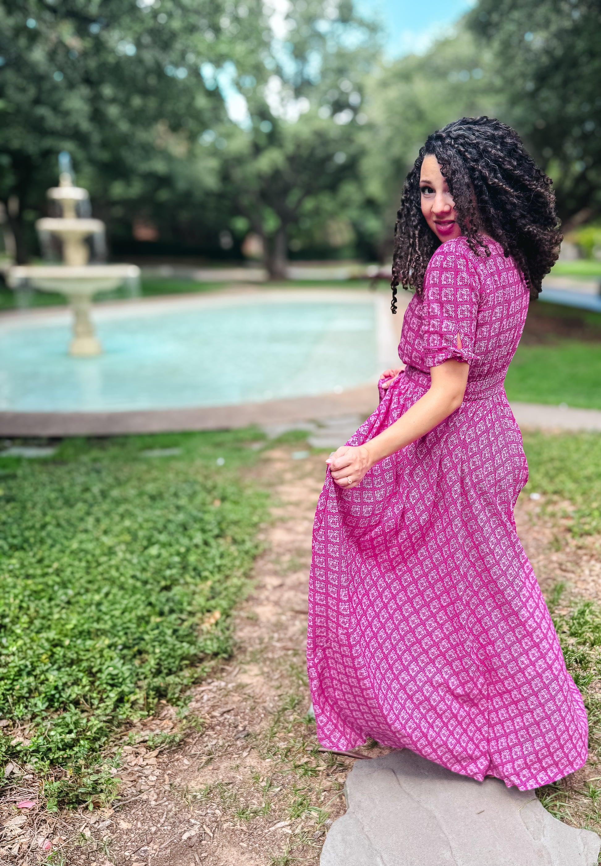 Woman in a pink modest nursing dress standing outdoors with a fountain and trees in the background