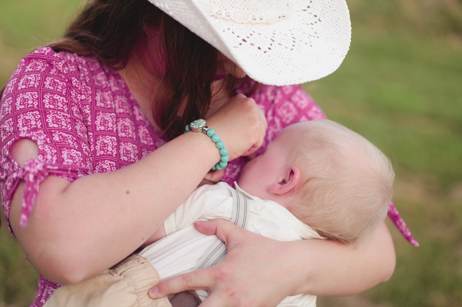 Woman in a pink modest nursing dress and white hat holding a baby outdoors.