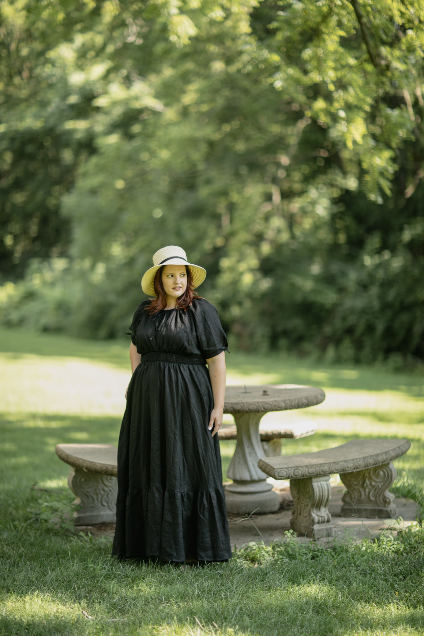 Woman in a black modest nursing dress and yellow hat standing in a park with stone benches and trees.