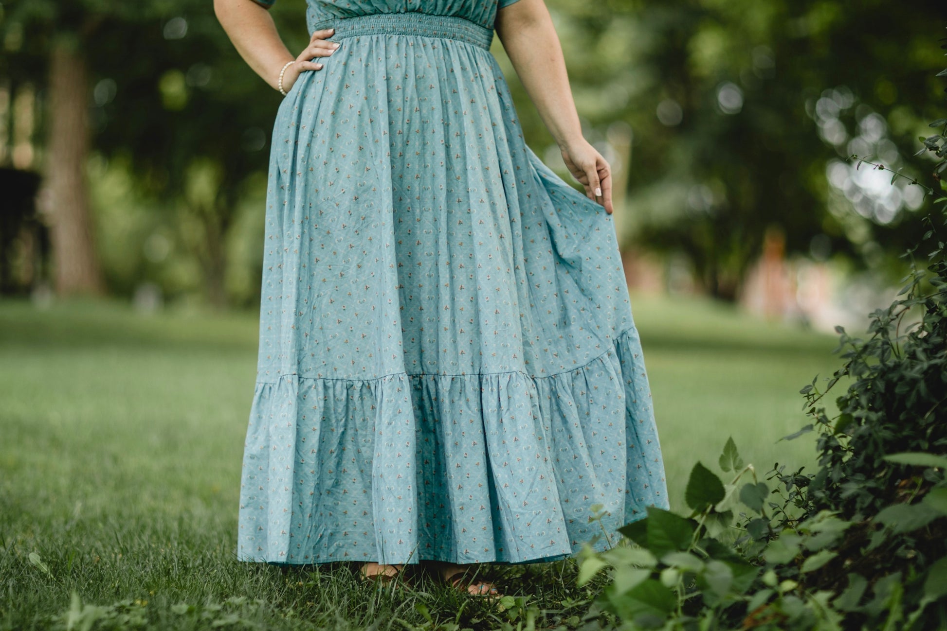 Woman wearing a long teal modest nursing dress standing in a grassy area with trees in the background