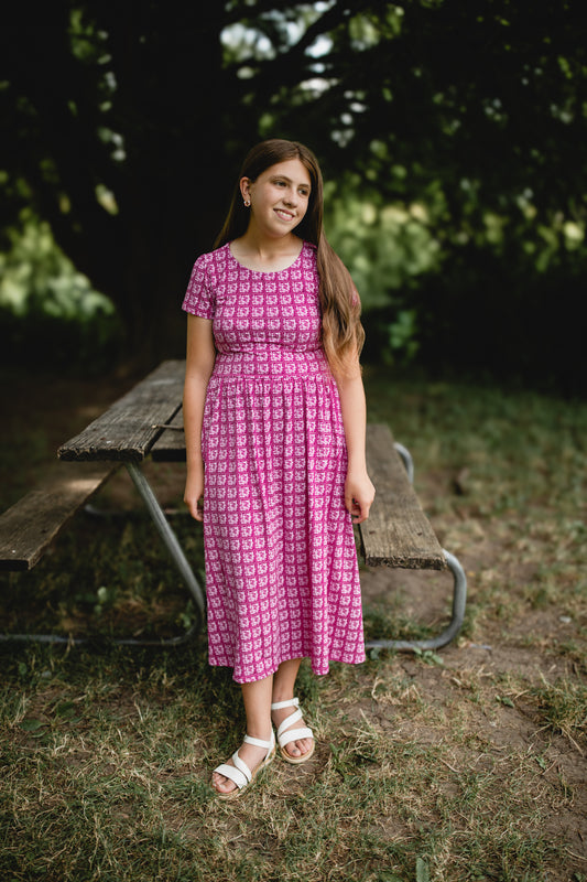 Young girl in a modest pink dress standing outdoors near a picnic table with greenery in the background