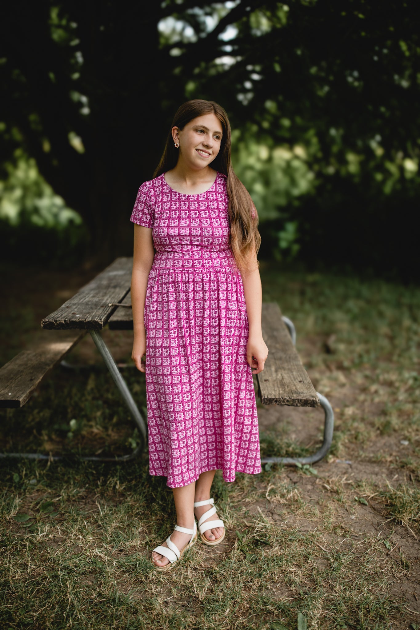 Young girl in a modest pink dress standing outdoors near a picnic table with greenery in the background
