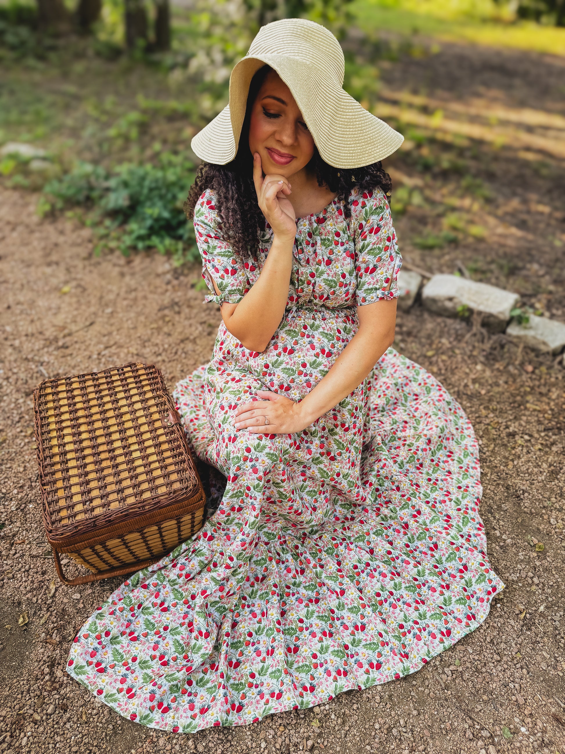 Woman in a floral modest nursing dress and sun hat sitting outdoors with a picnic basket.