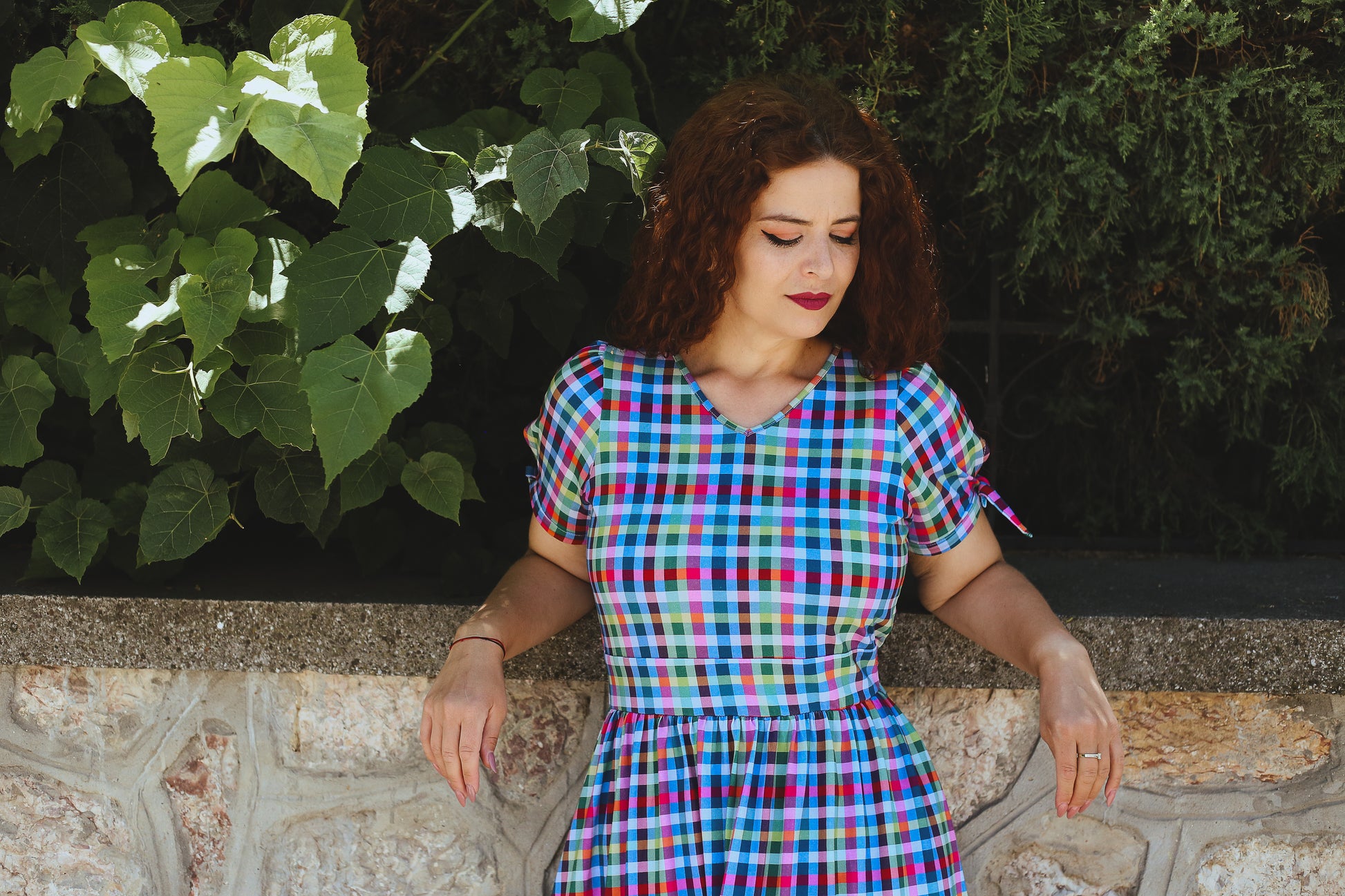 Woman in a colorful checkered modest nursing dress sitting against a stone wall with greenery.