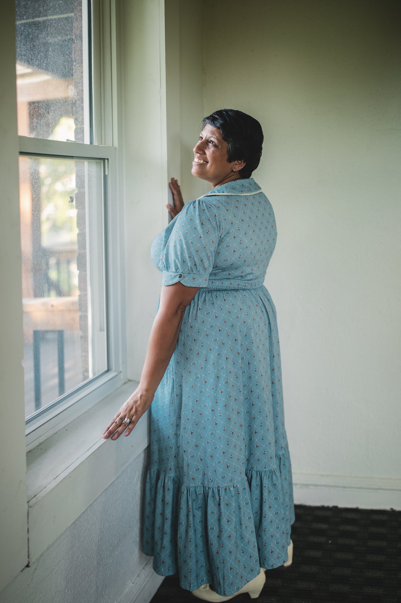 Woman in a blue modest nursing dress standing by a window, looking out.