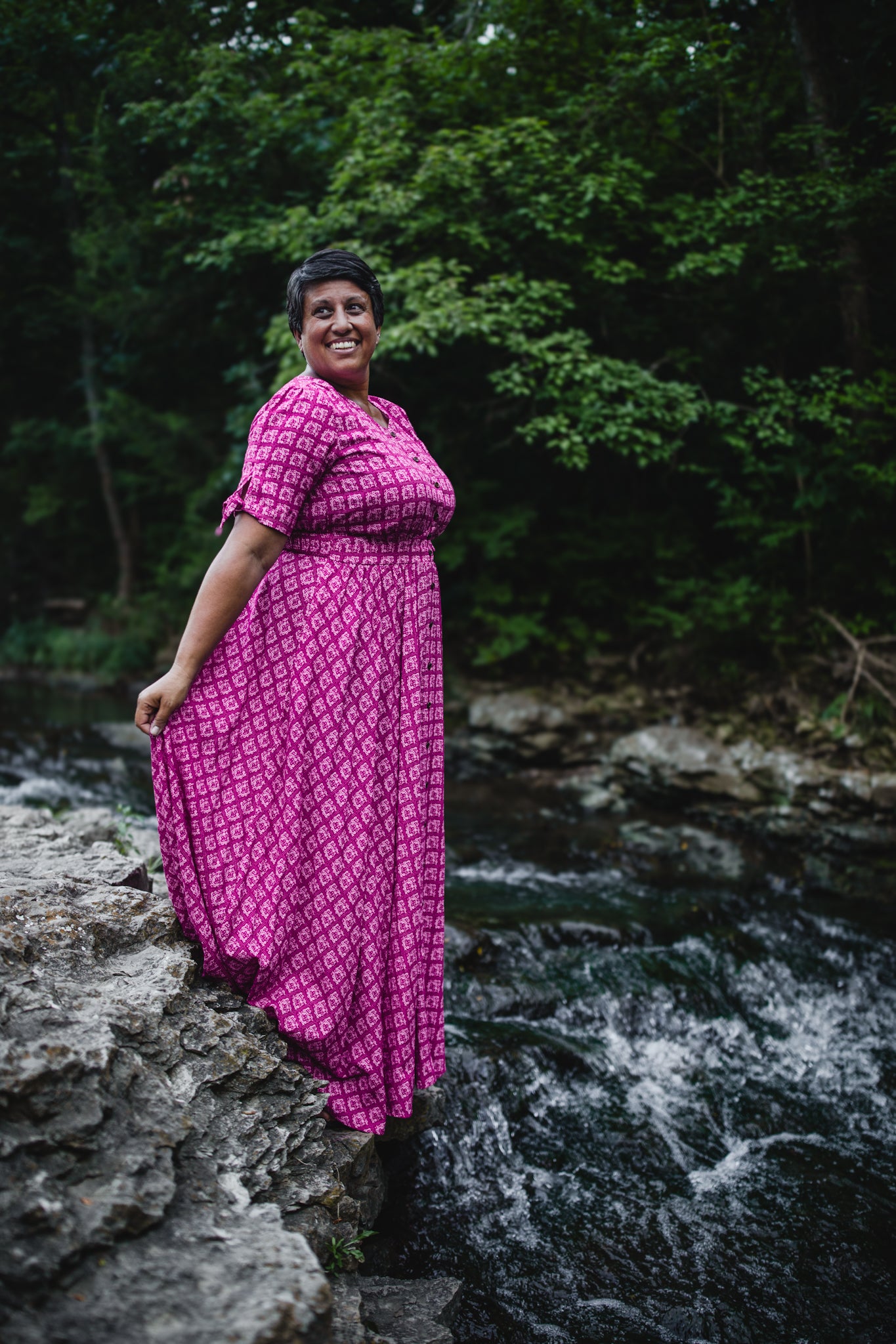 Woman in a pink modest nursing dress standing by a stream with greenery in the background