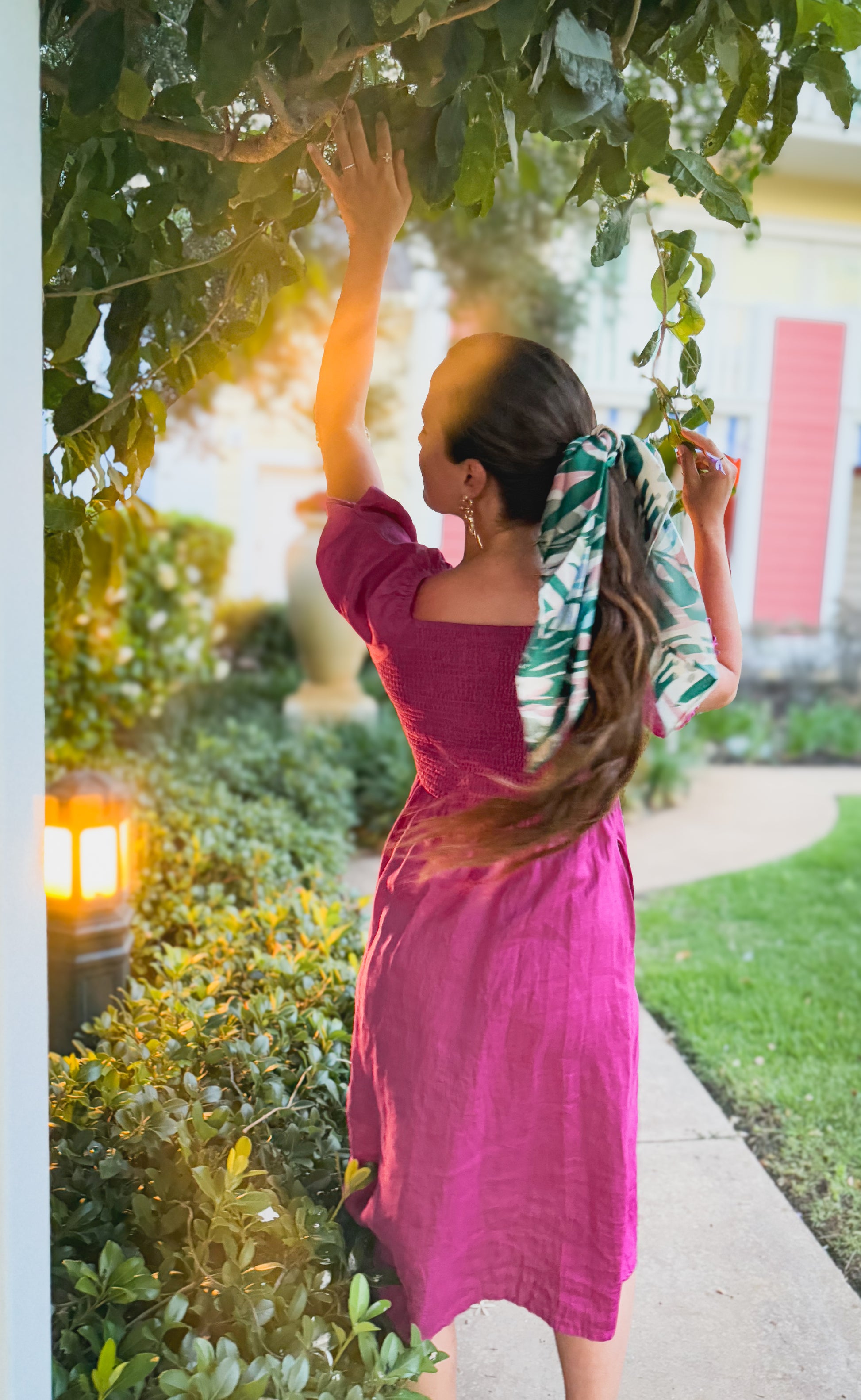 Woman in a pink modest nursing dress with a green and white patterned scarf, standing outdoors with trees and a house in the background.