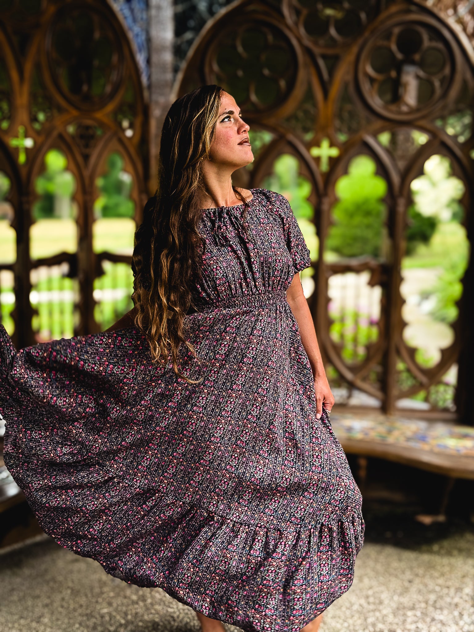 Woman in a floral modest nursing dress standing in front of a decorative stone wall with a garden view.