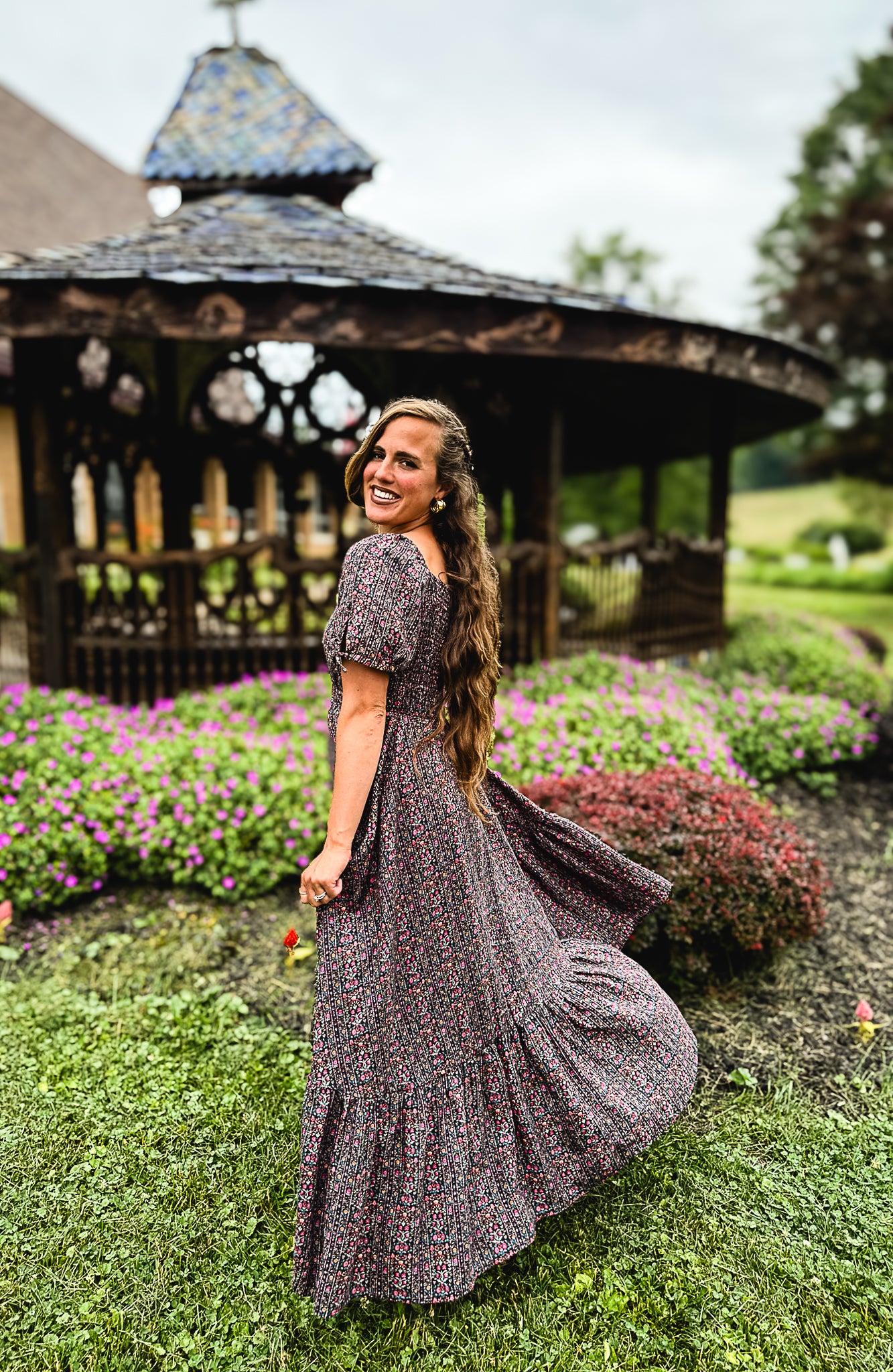 Woman in a floral modest nursing dress standing in a garden with a gazebo in the background