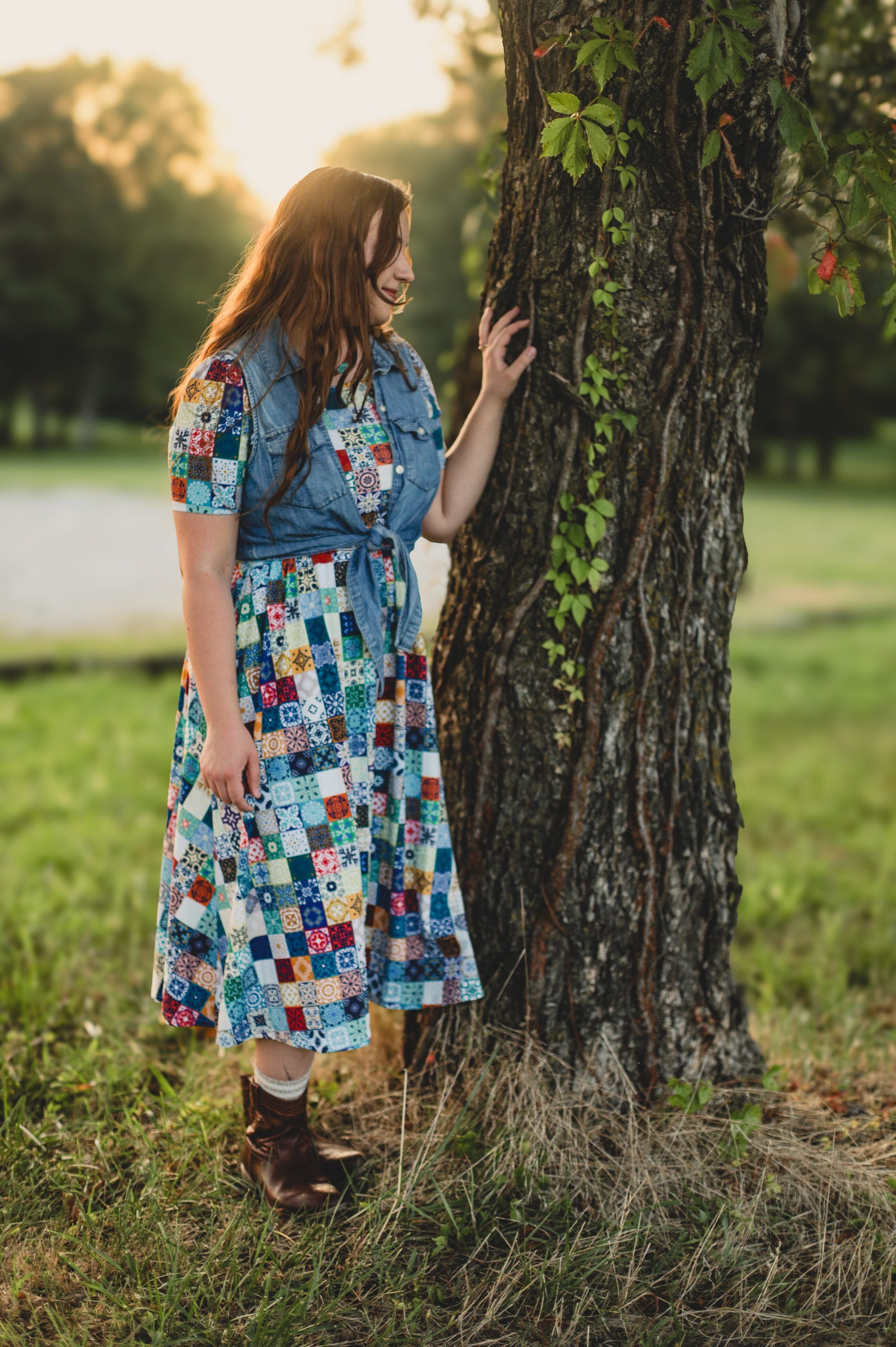 Woman in a colorful modest nursing dress standing next to a tree in a park at sunset.
