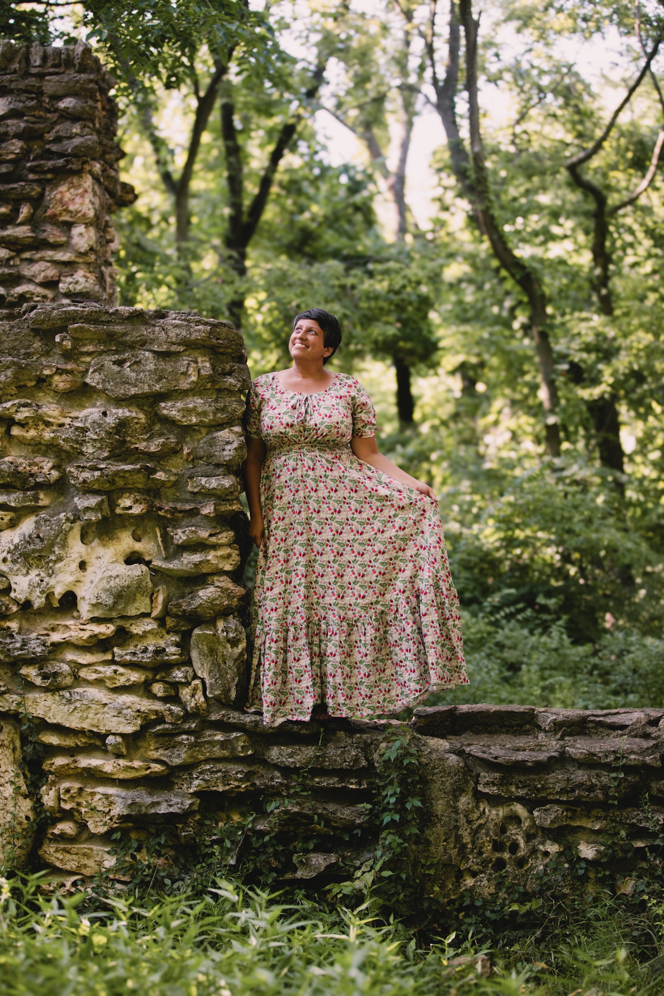 Woman in a floral modest nursing dress standing against a stone wall in a forest setting