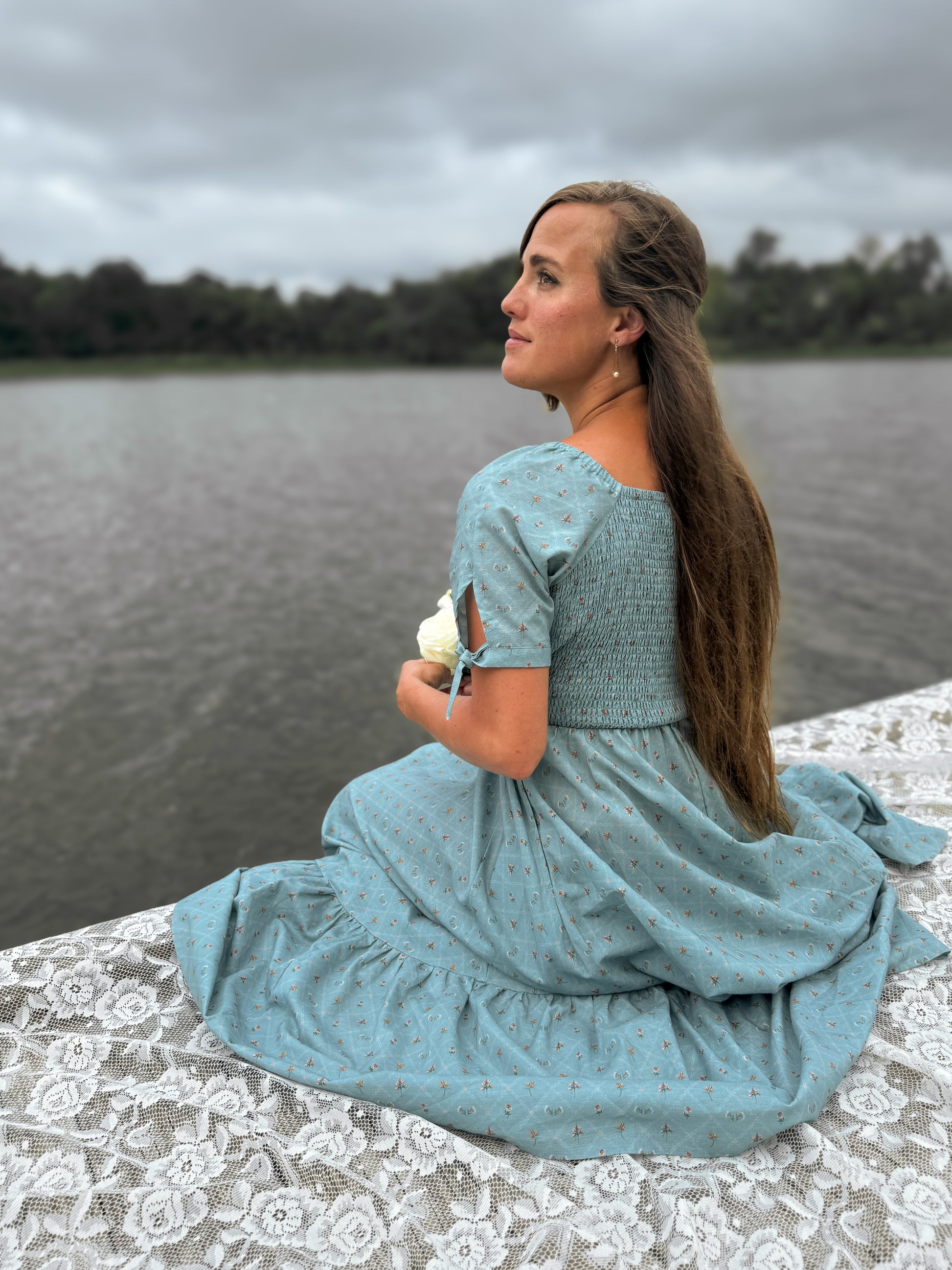 Woman in a blue modest nursing dress sitting by a lake on a cloudy day