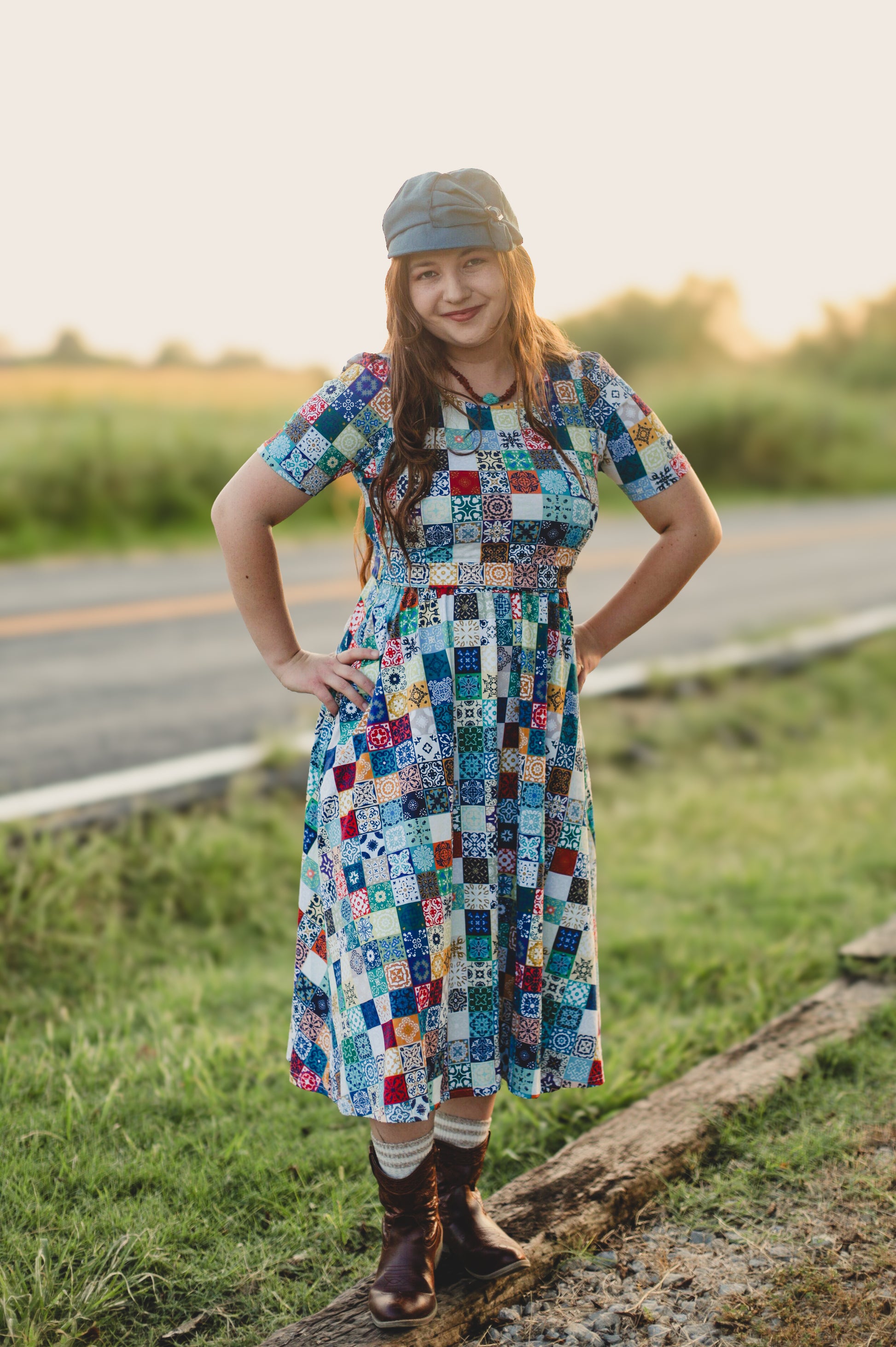 Woman in a colorful patchwork modest nursing dress standing on a grassy roadside.