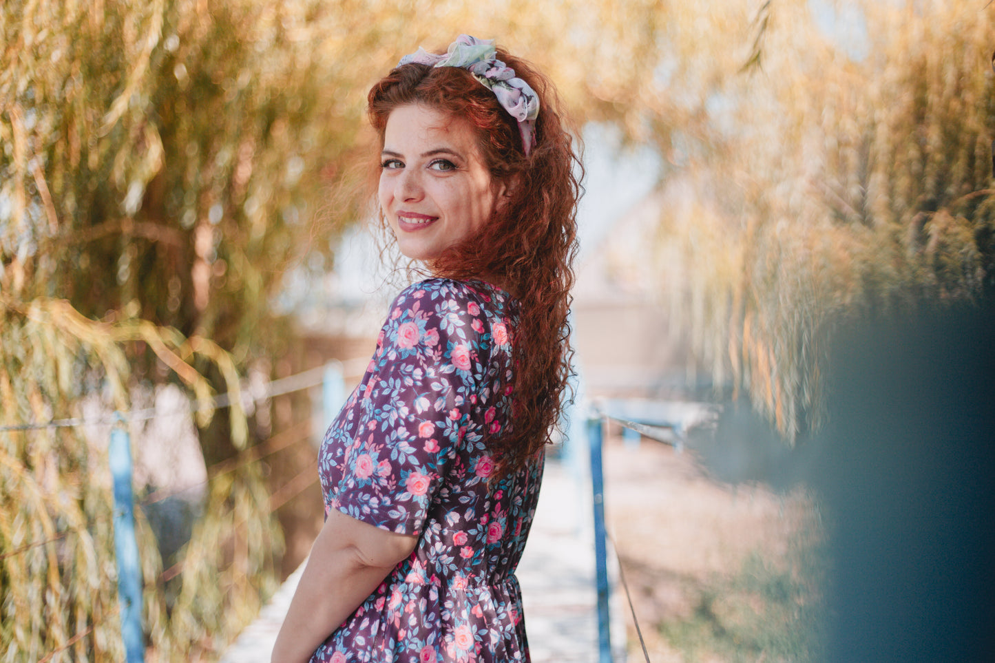 Woman with long red hair wearing a modest nursing floral dress and headband, standing outdoors by a body of water.