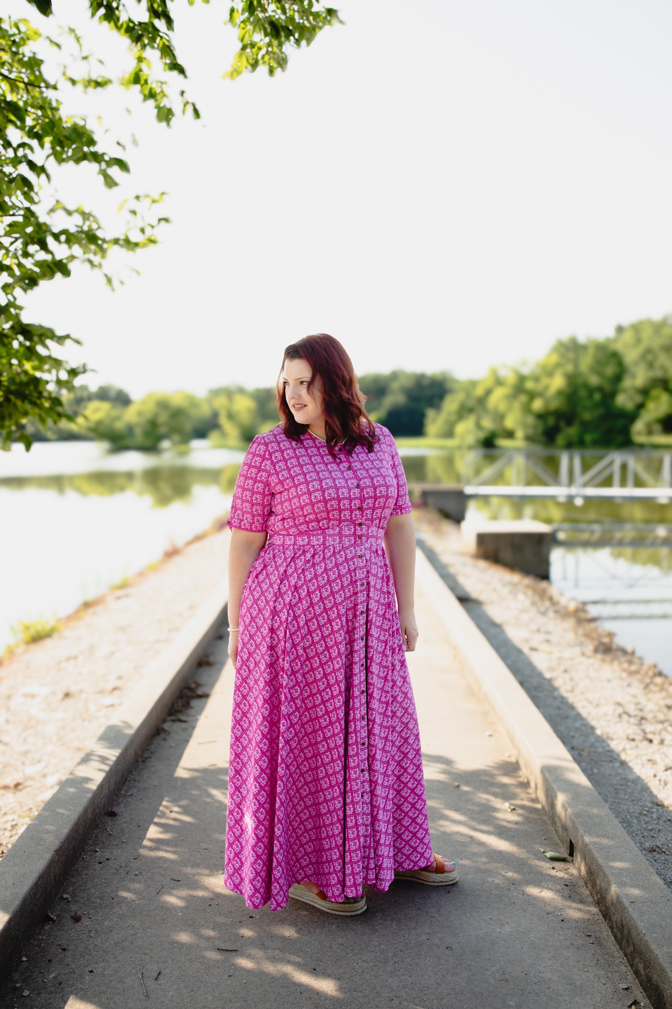 Woman in a pink modest nursing dress standing on a path by a river with trees in the background