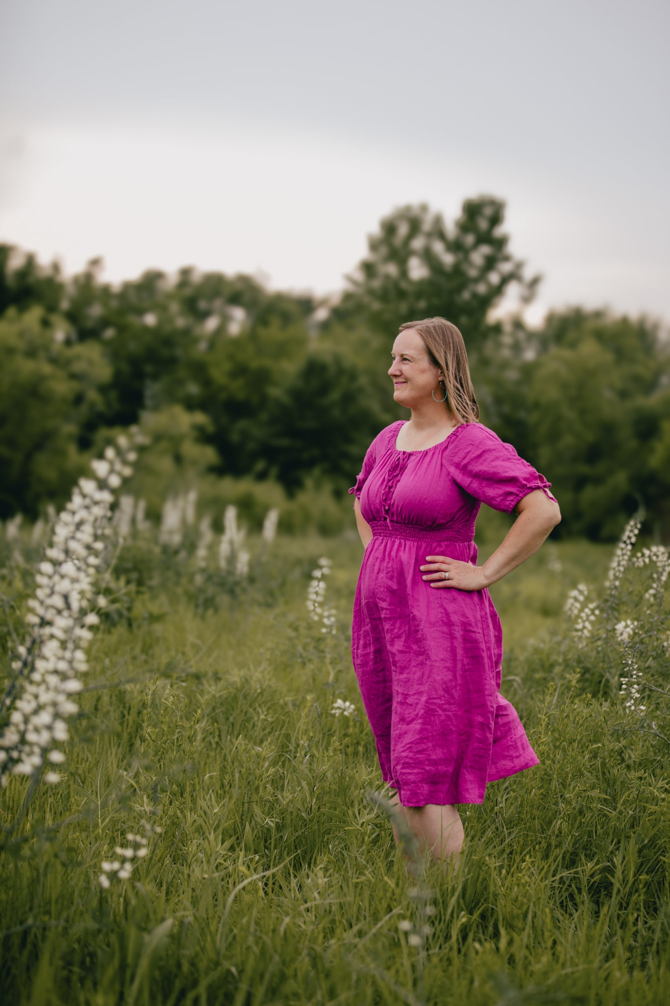 Woman in a pink modest nursing dress standing in a field with greenery and white flowers.