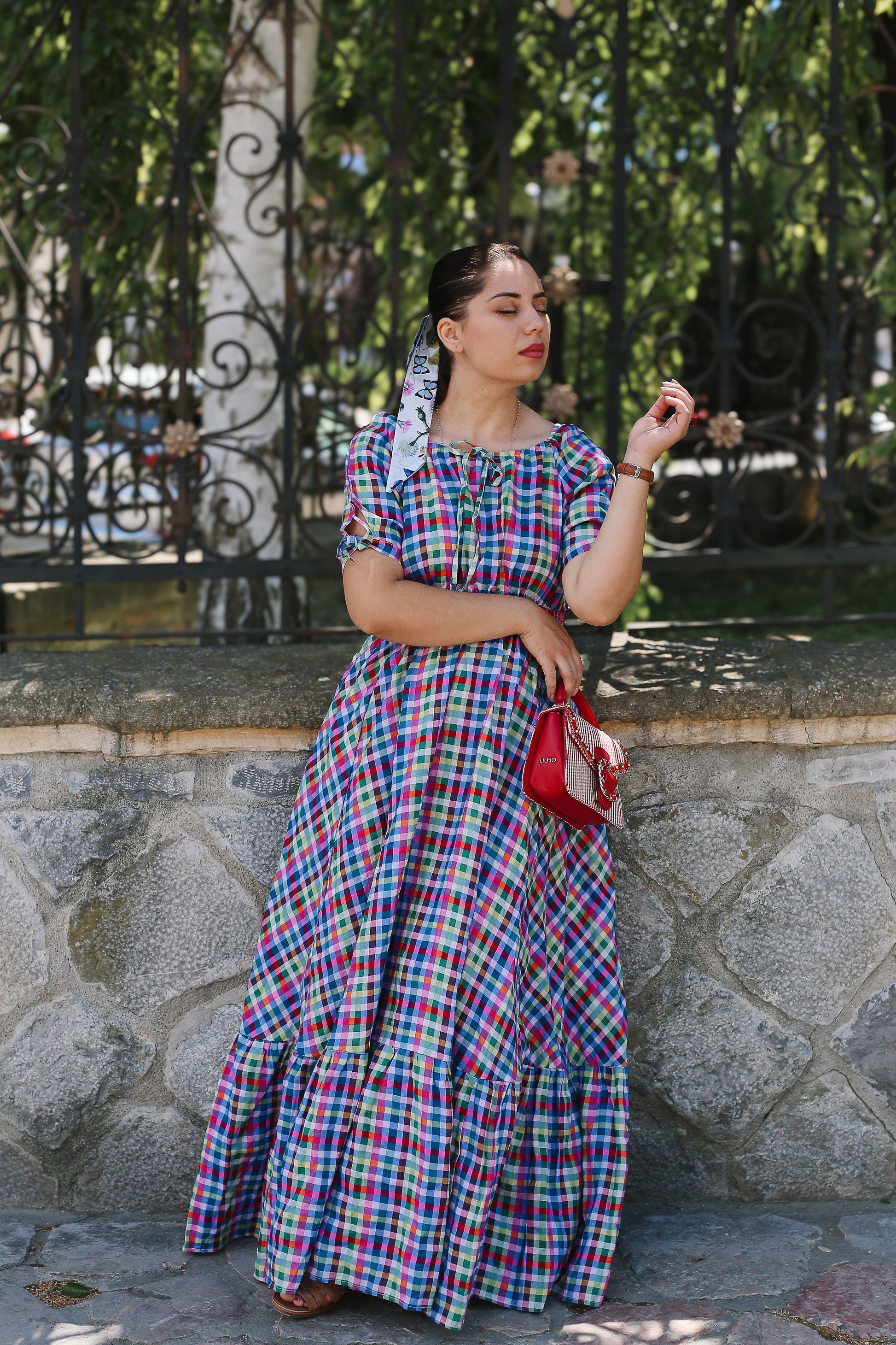 Woman in a colorful plaid modest nursing dress standing outdoors with a stone wall and trees in the background.