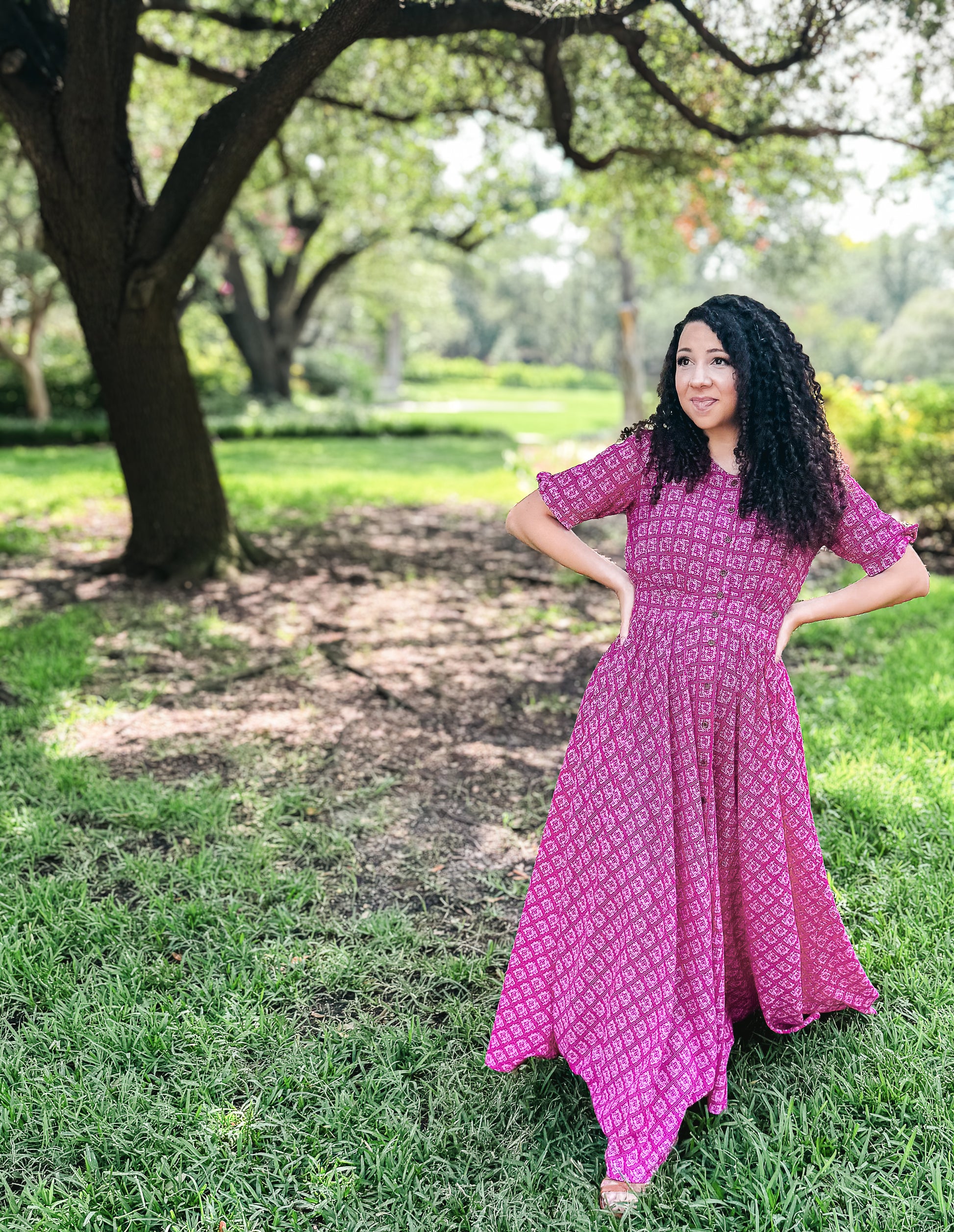 Woman in a pink modest nursing dress standing in a park with trees and grass.