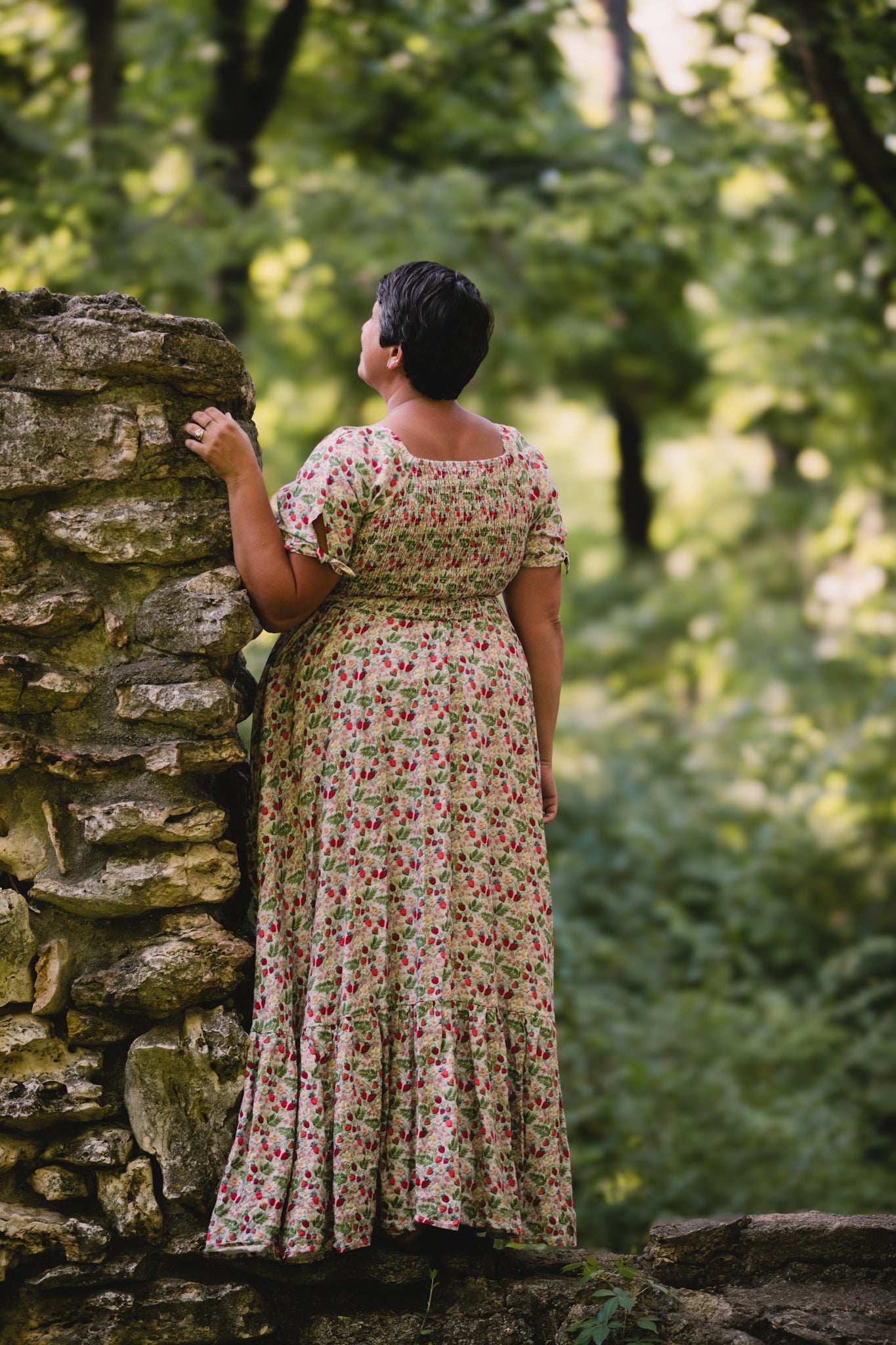 Woman in a floral modest nursing dress standing by a stone wall in a forest