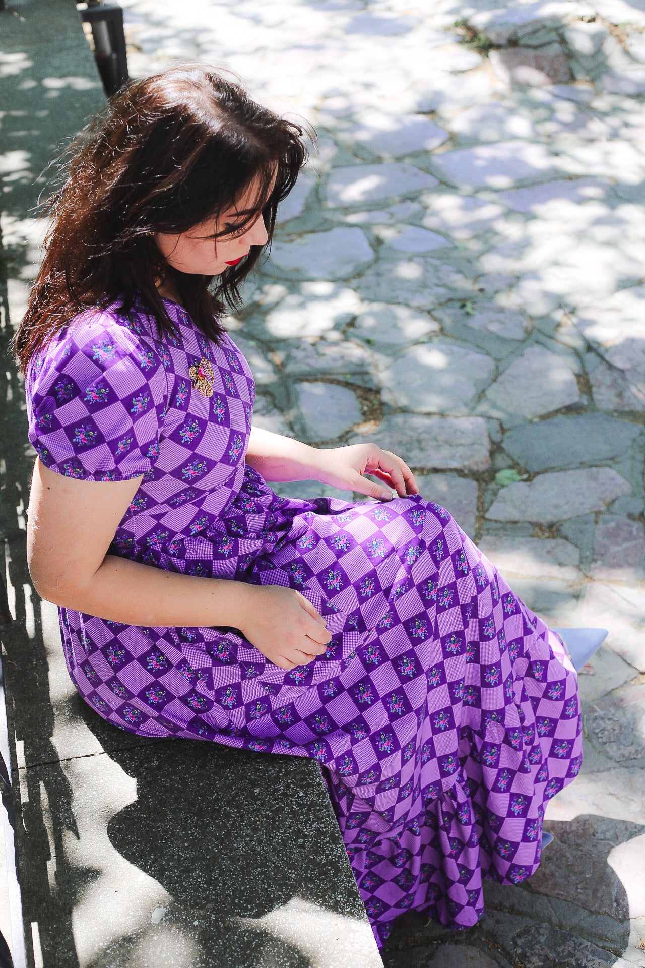 Woman in a purple patterned modest nursing dress sitting outdoors on a stone path.