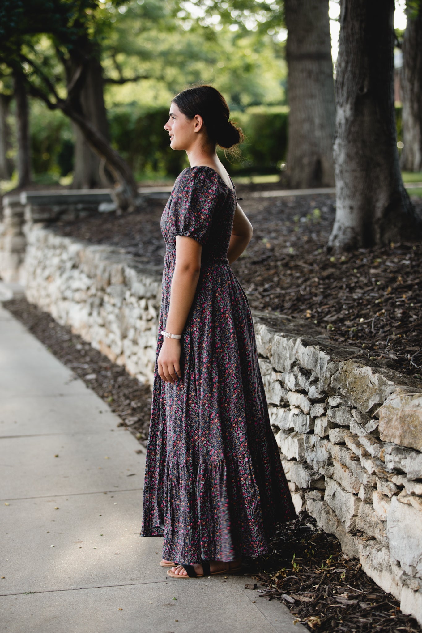 Woman in a long, dark floral modest nursing dress standing on a sidewalk with trees and stone wall in the background