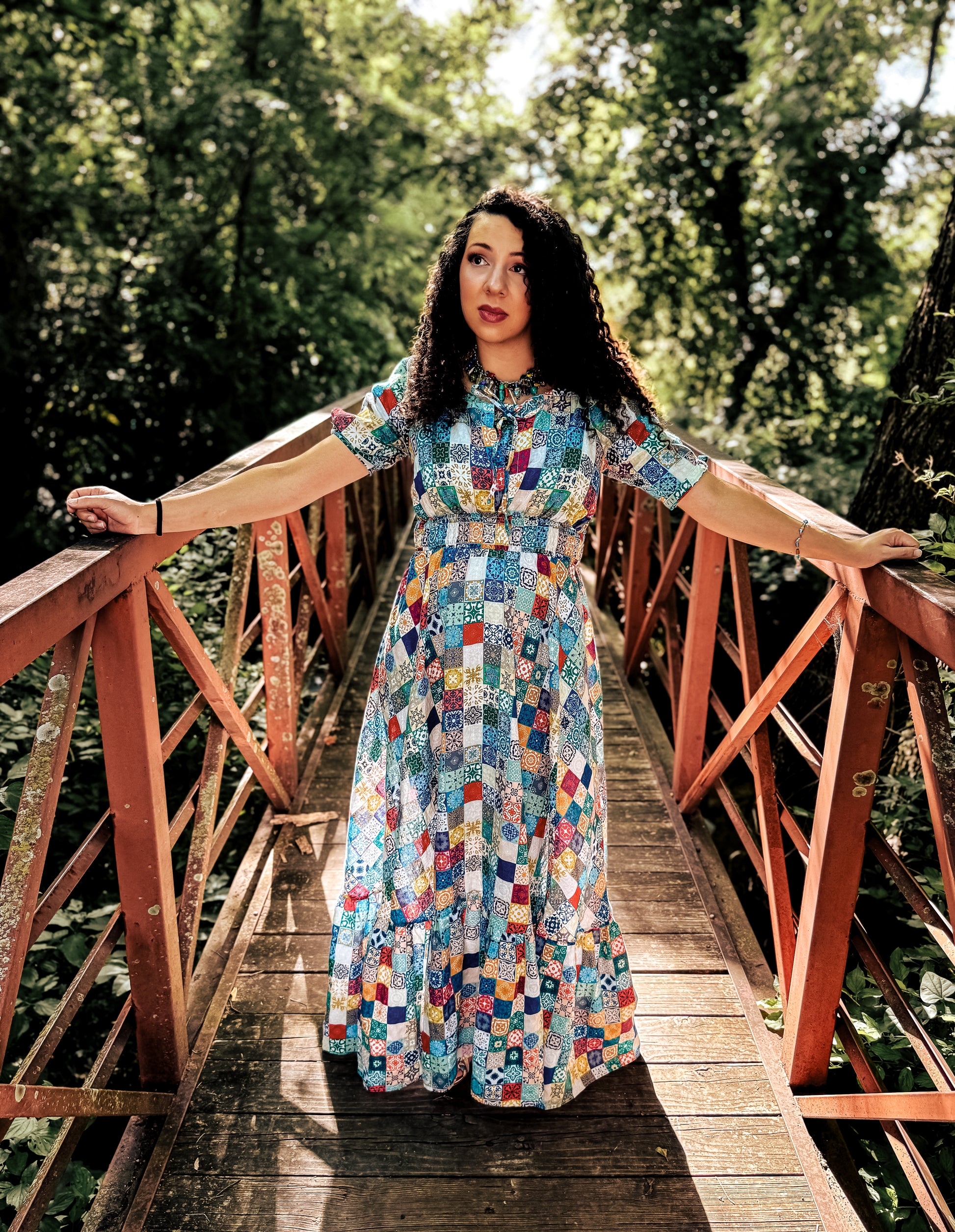 Woman in a colorful modest nursing dress standing on a wooden bridge with trees in the background