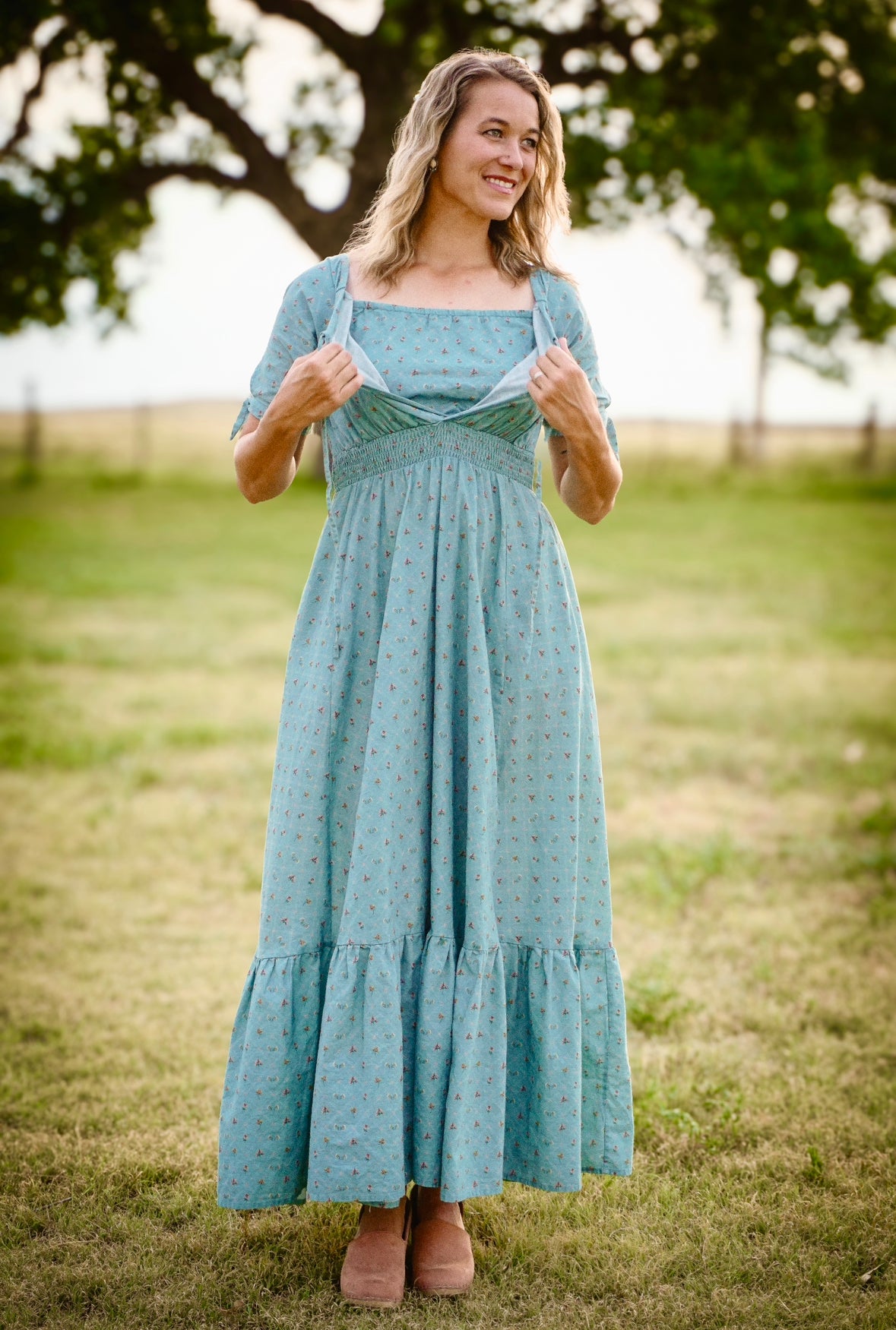 Woman wearing a teal modest nursing dress standing in a grassy field with trees in the background