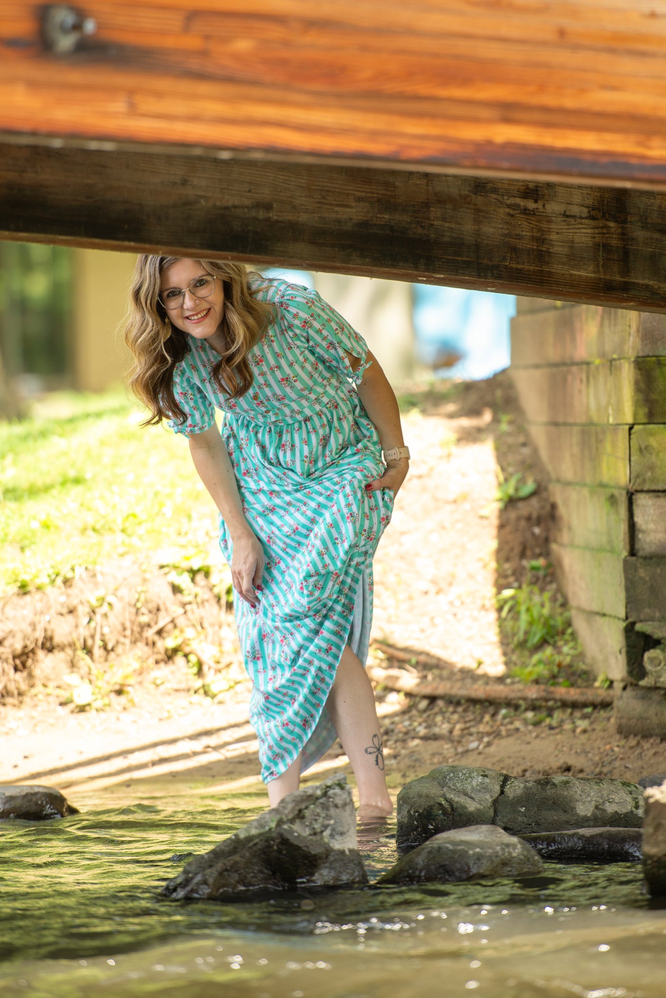 Woman in a blue modest nursing dress standing under a wooden bridge by a stream.