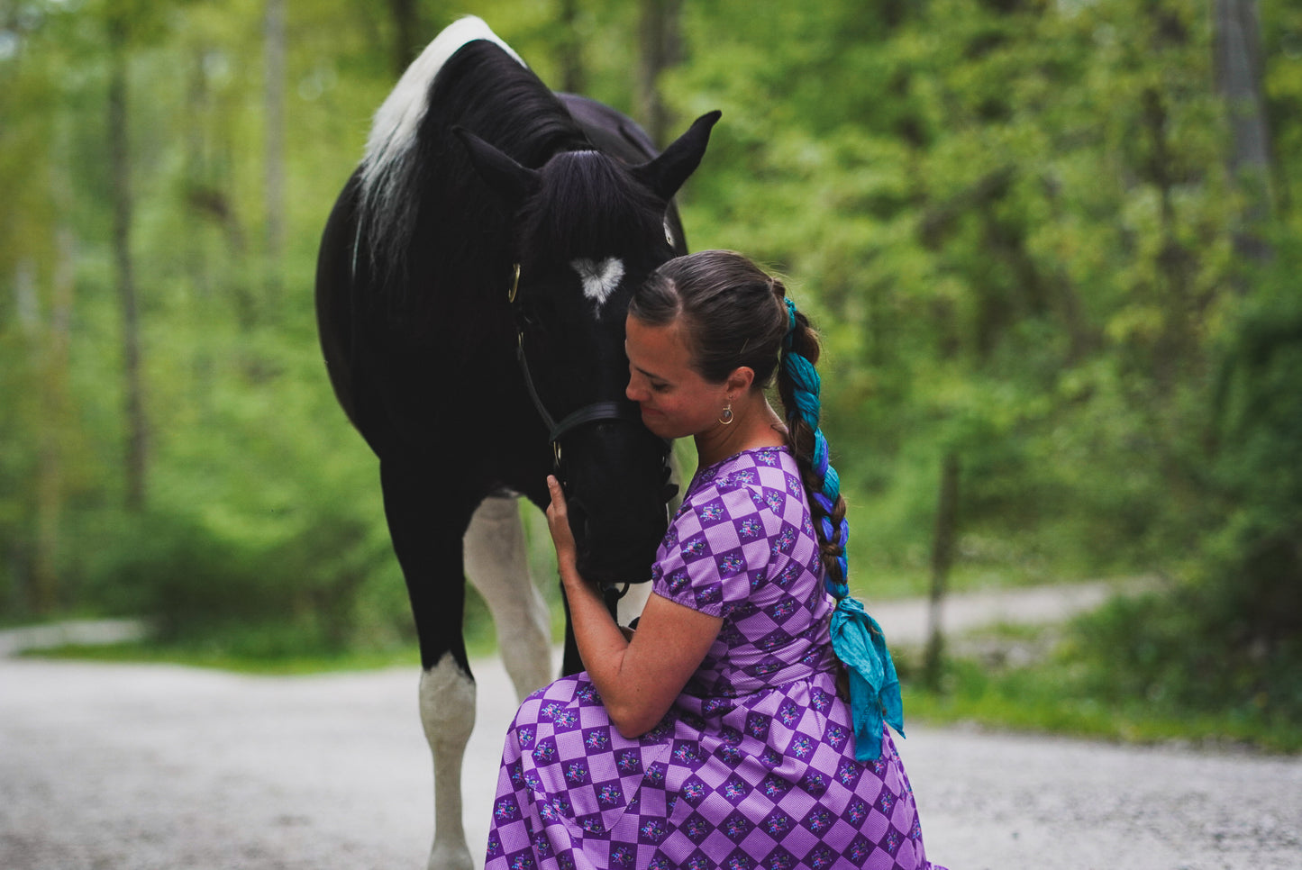 Woman in a purple checkered modest nursing dress petting a black horse on a path surrounded by trees.