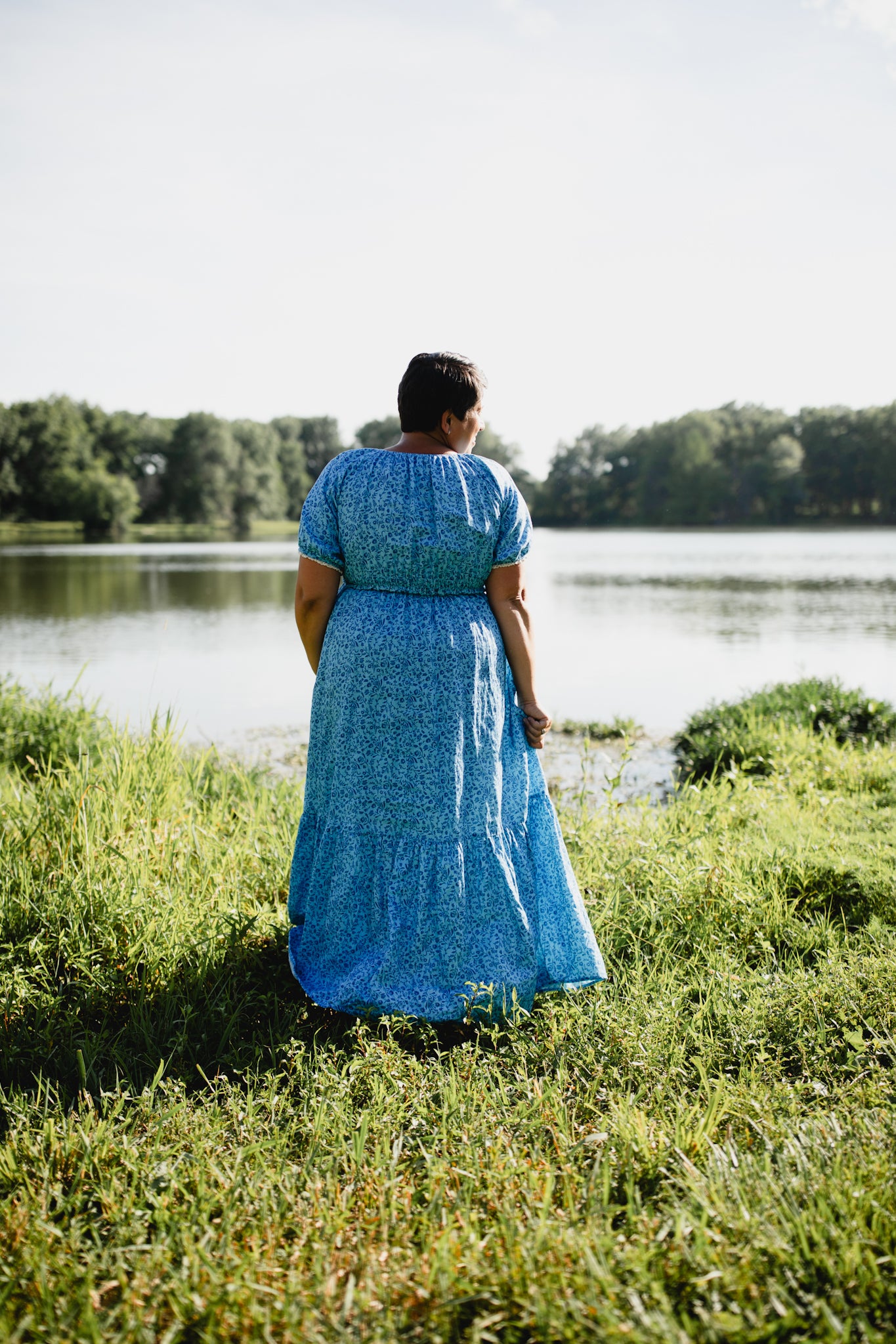 Woman in a blue modest nursing dress standing by a lake with trees in the background