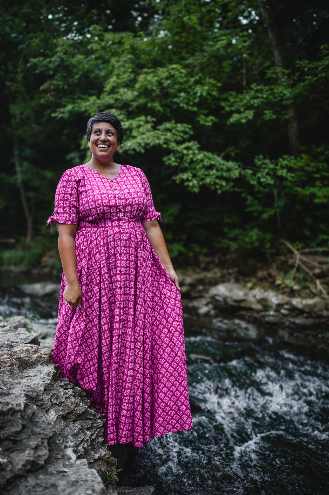 Woman in a pink modest nursing dress standing by a stream with greenery in the background