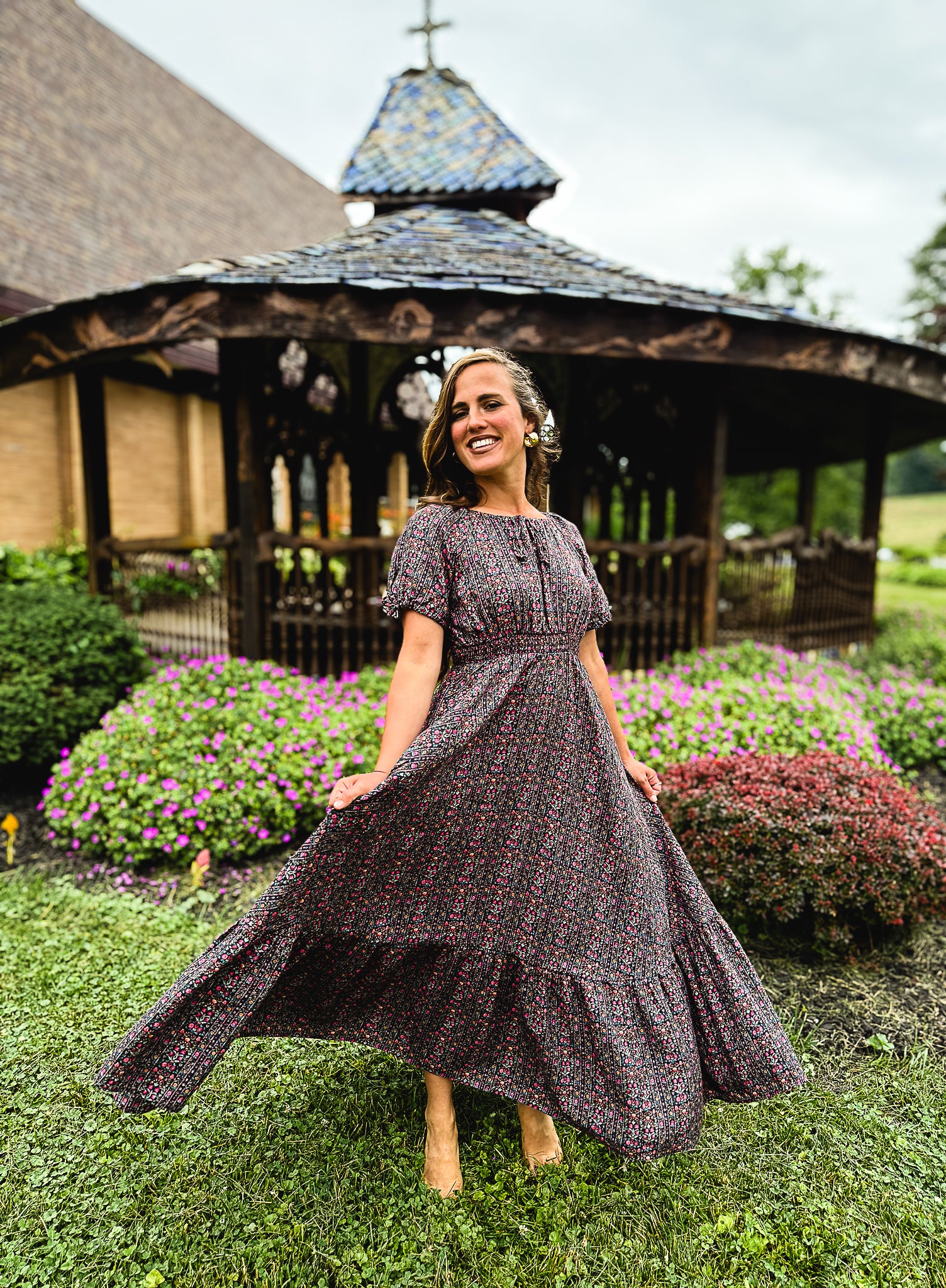 Woman in a floral modest nursing dress standing outdoors with a gazebo and flowers in the background