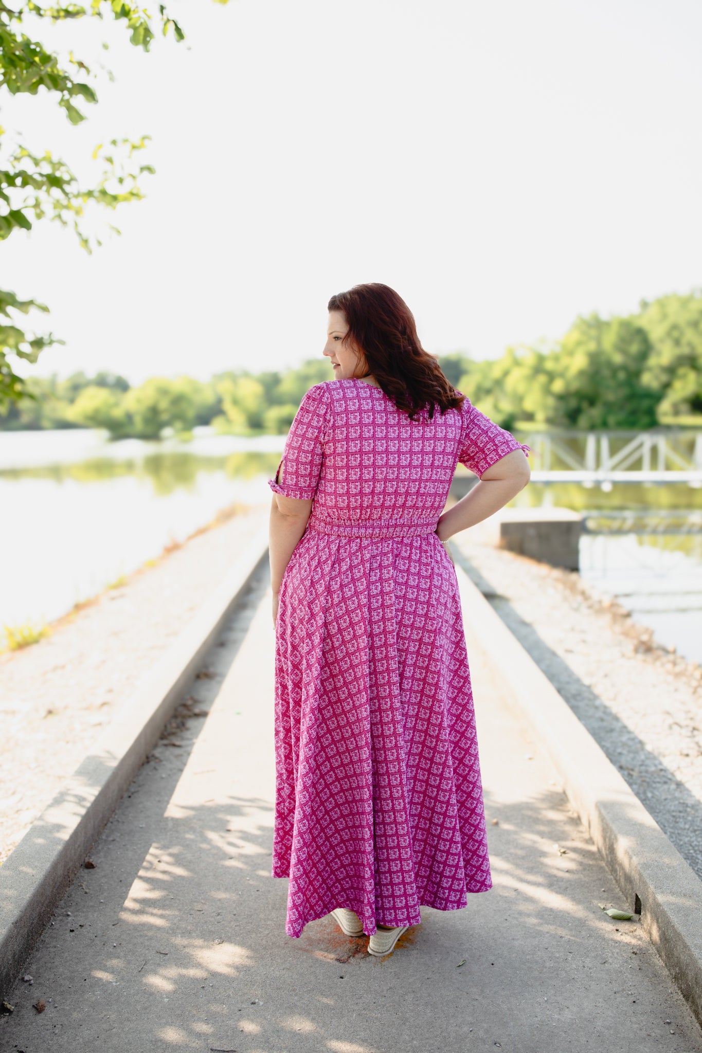 Woman in a pink modest nursing dress standing by a lake with trees in the background
