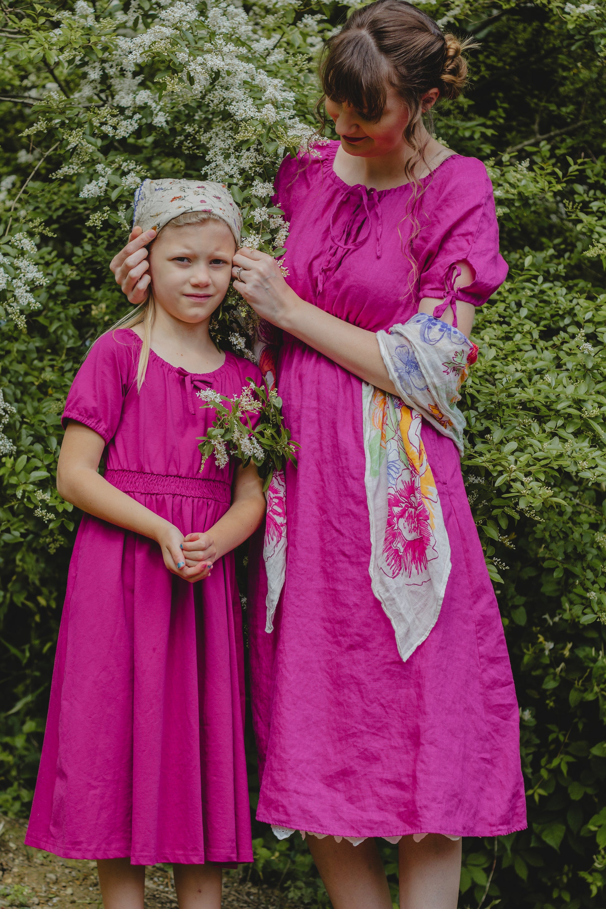 Mother and daughter in bright pink modest dresses standing outdoors with greenery in the background