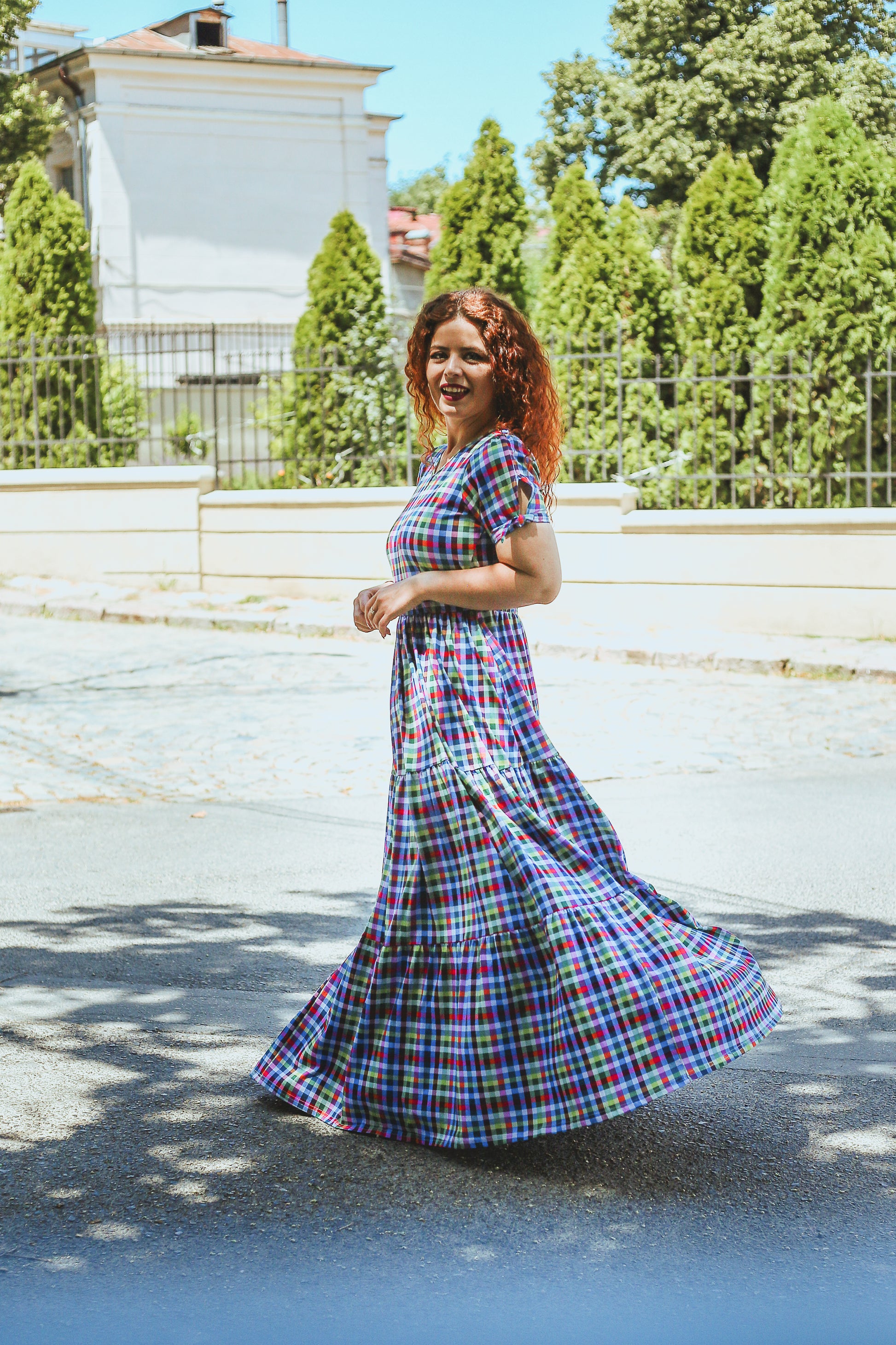 Woman in a plaid modest nursing dress standing outdoors with trees and buildings in the background