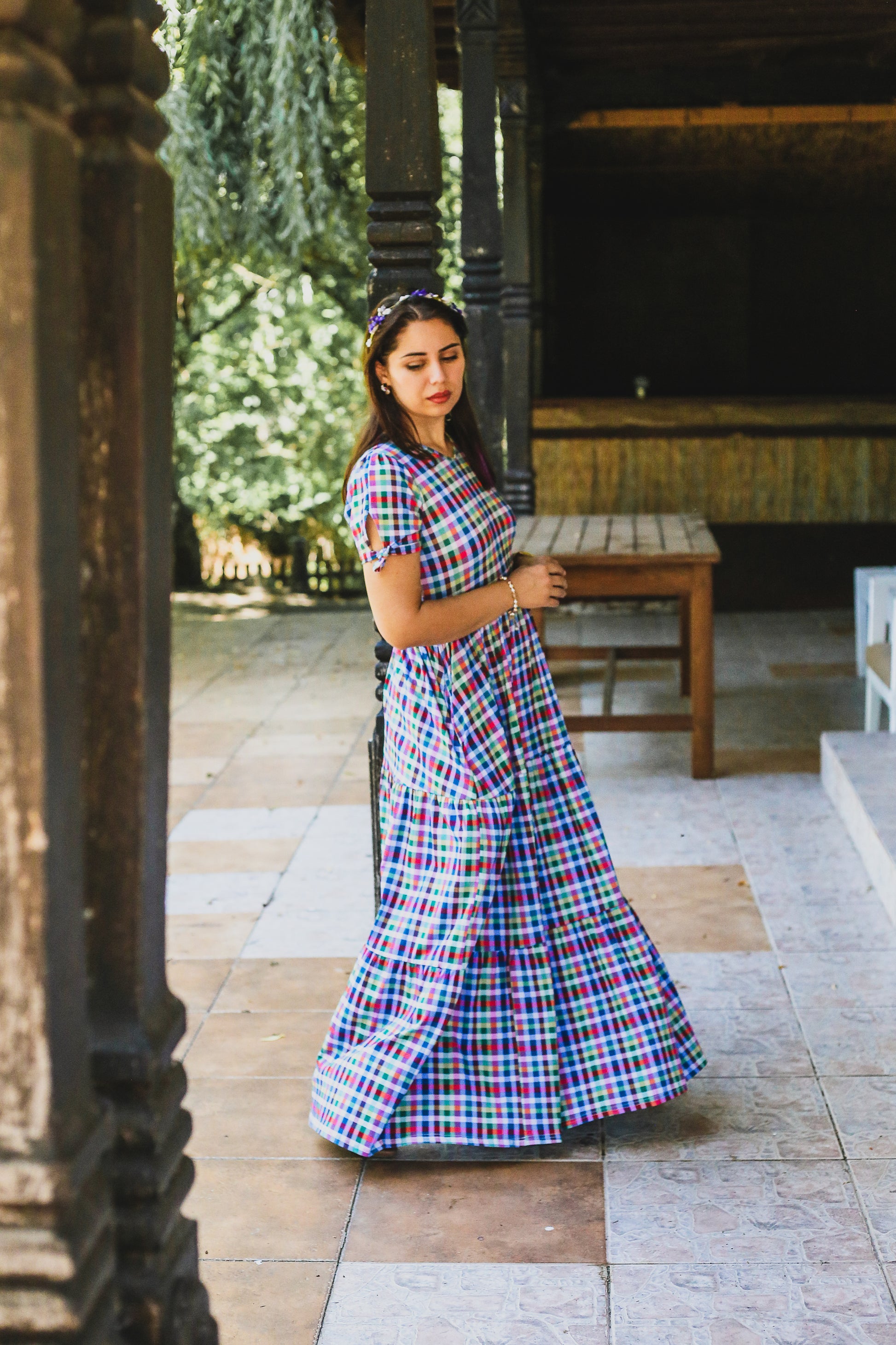 Woman in a colorful plaid modest nursing dress standing in an outdoor setting with stone flooring and wooden furniture.