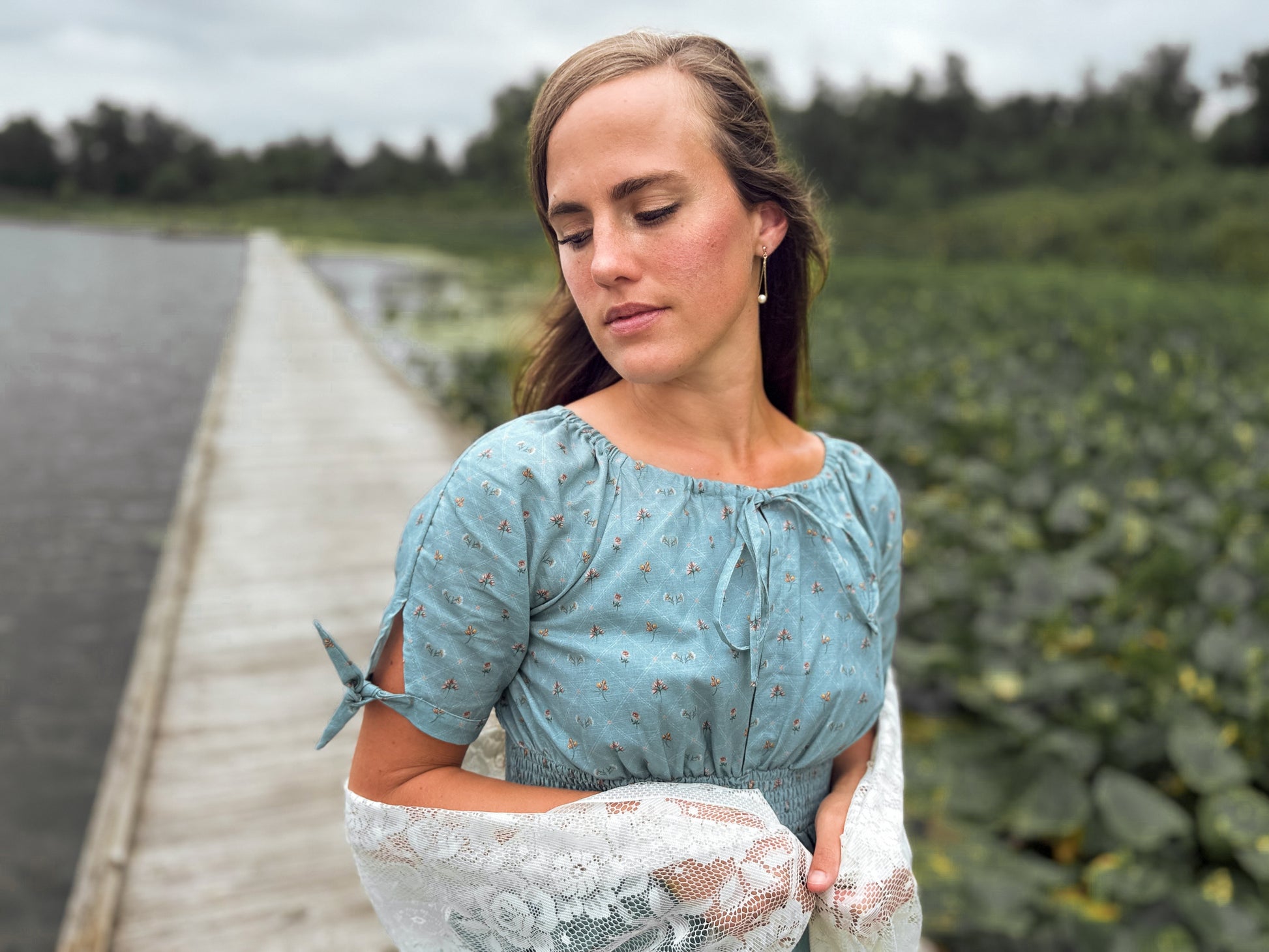 Woman in a blue modest nursing dress standing on a wooden dock with a field and water in the background