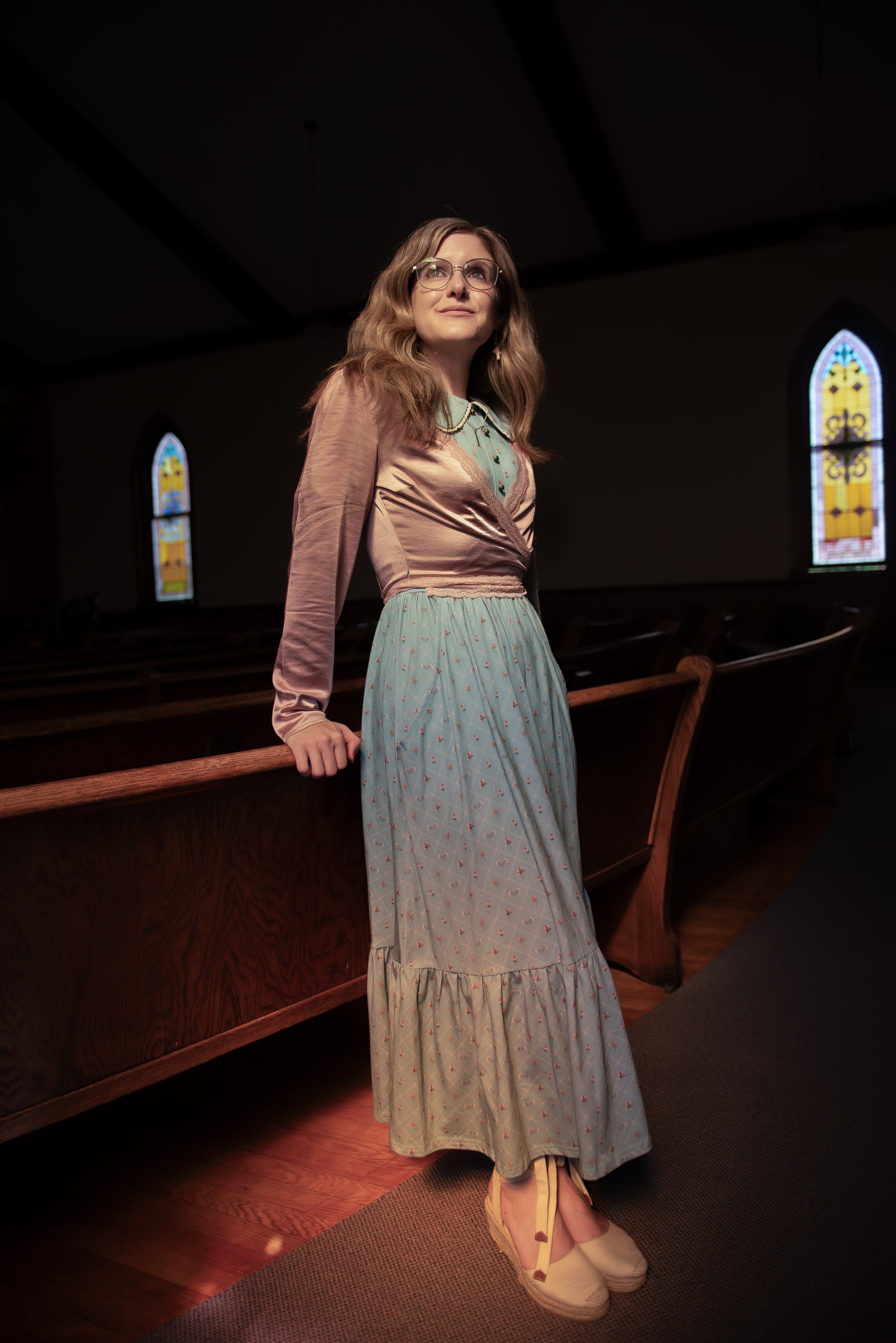 Woman in a long modest nursing dress standing in a dimly lit room with stained glass windows.