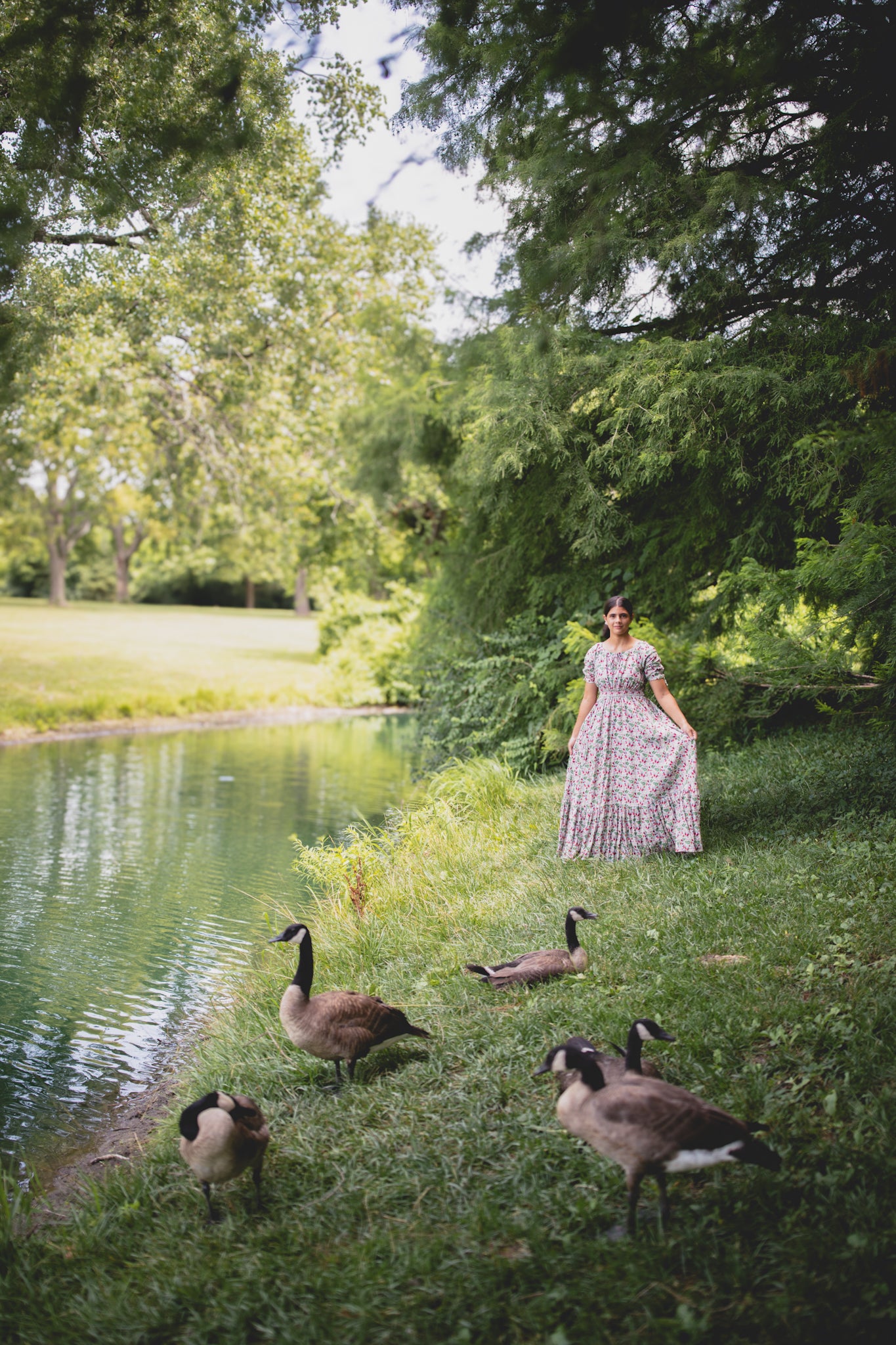 Woman in a modest nursing dress standing by a pond with geese in a park-like setting