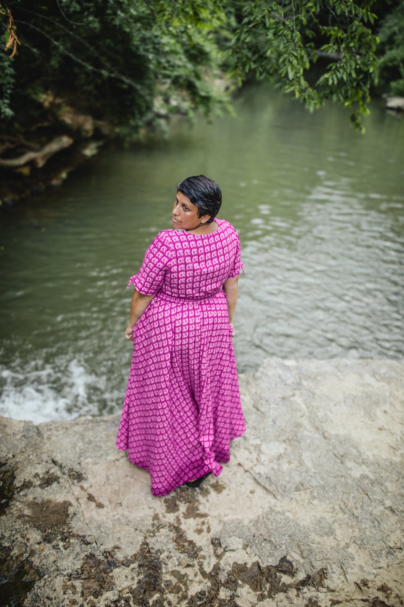 Woman in a pink modest nursing dress standing by a body of water with greenery in the background