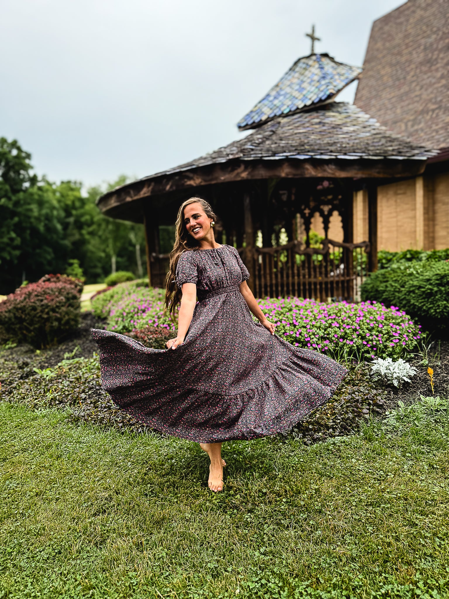 Woman in a flowing modest nursing dress standing in a garden with a gazebo in the background