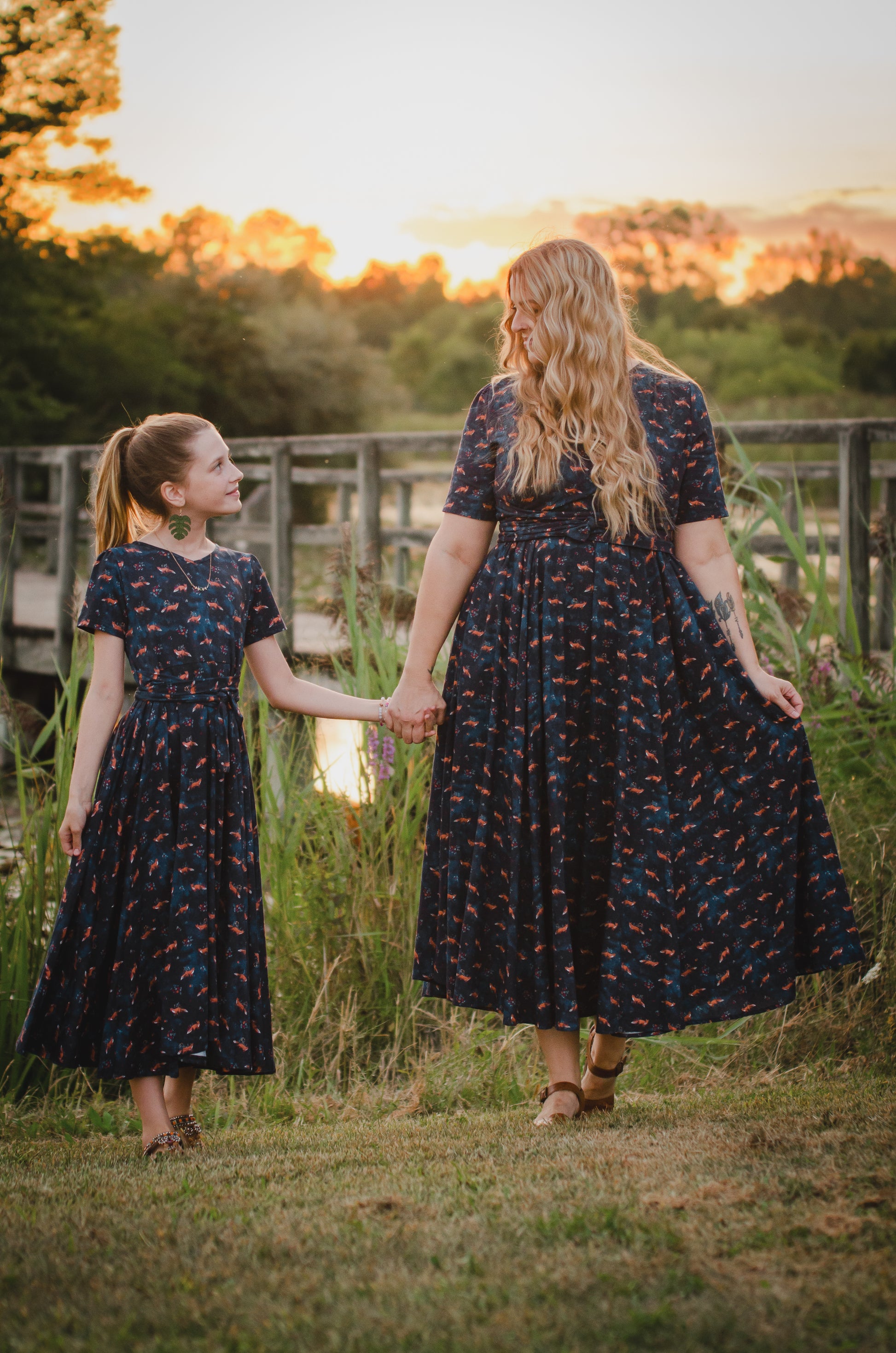 Two women in matching modest, nursing-friendly dark floral dresses walking hand-in-hand at sunset