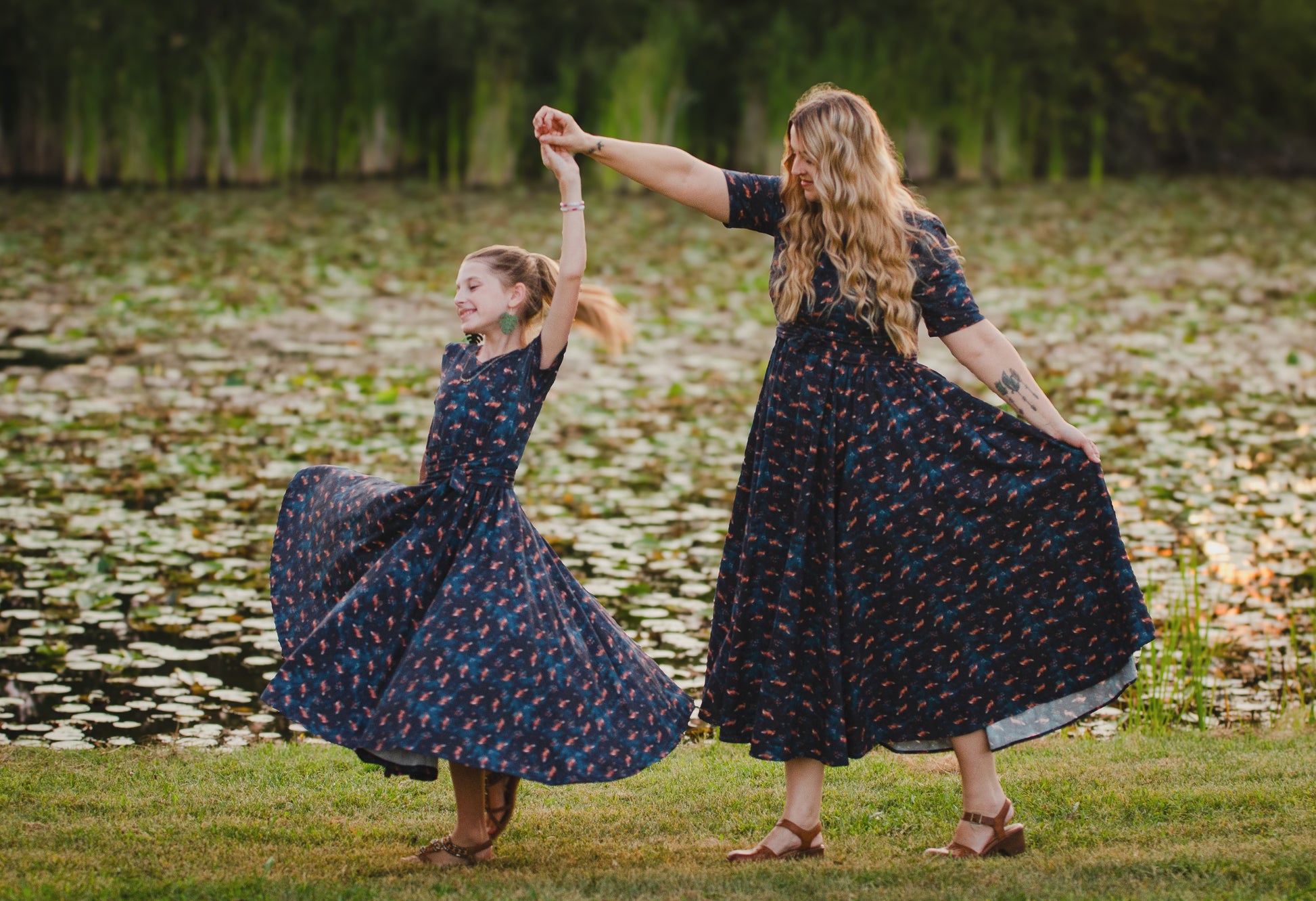 Two women in matching modest, nursing-friendly dark floral dresses dancing outdoors by a pond
