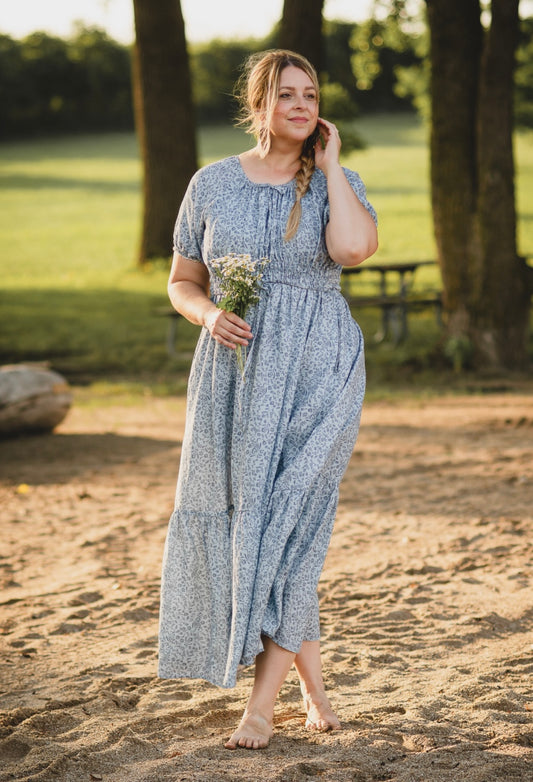 Woman in a modest blue nursing dress standing on a sandy path with trees and grass in the background
