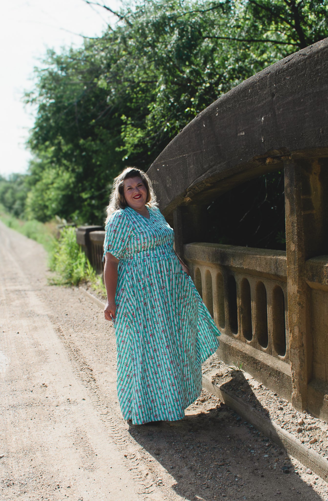 Woman in a modest nursing dress standing next to an old stone bridge on a dirt road.