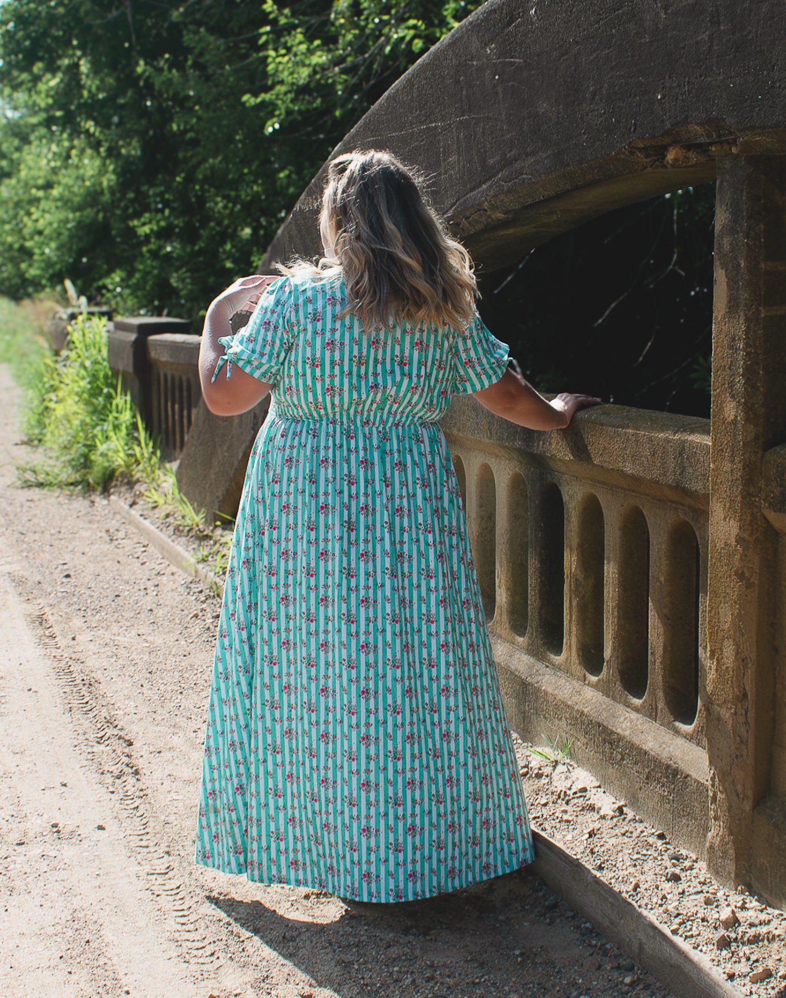 Woman in a patterned modest nursing dress standing on a bridge over a stream with trees in the background