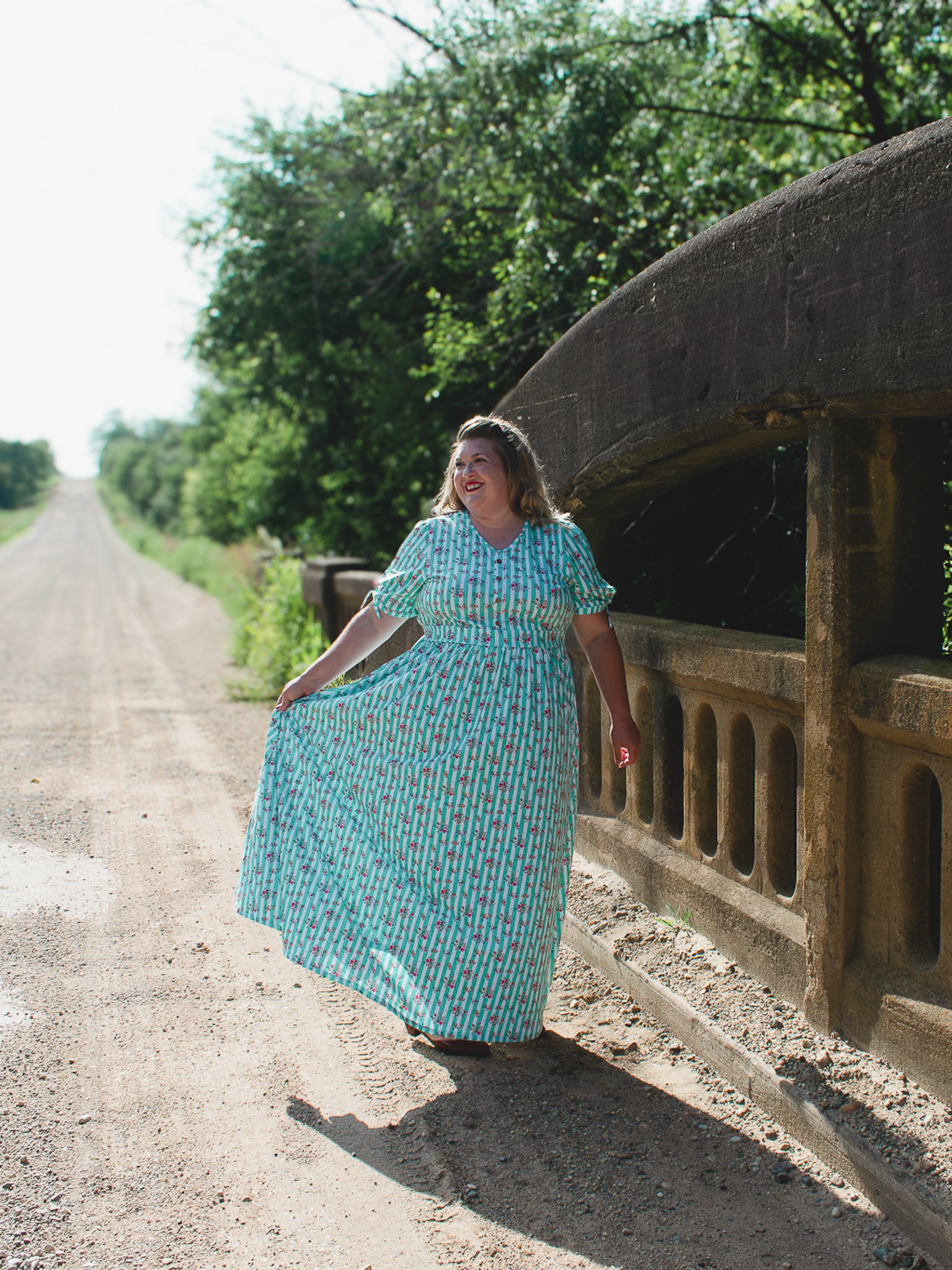 Woman in a blue modest nursing dress standing on a dirt road next to a large concrete structure.