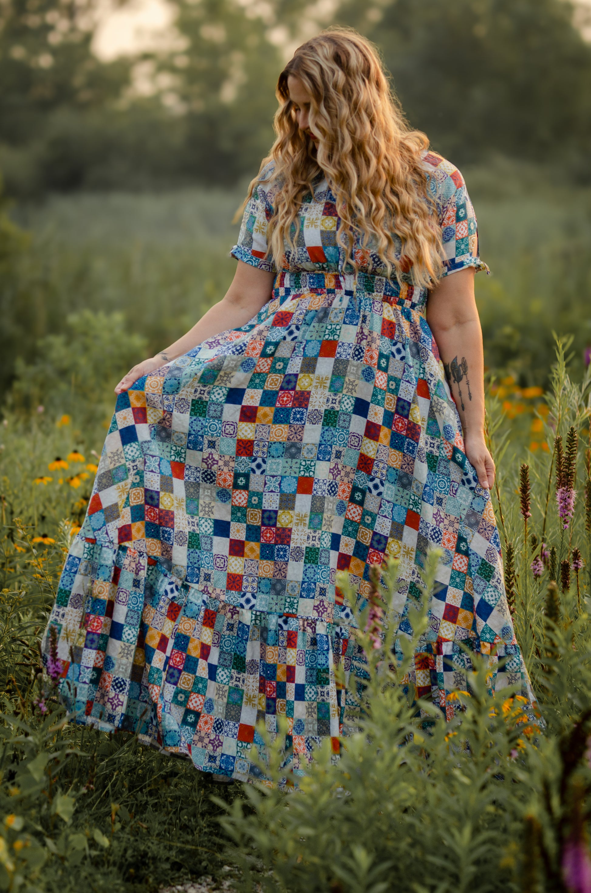 Woman in a colorful checkered modest nursing dress standing in a field with greenery and flowers.