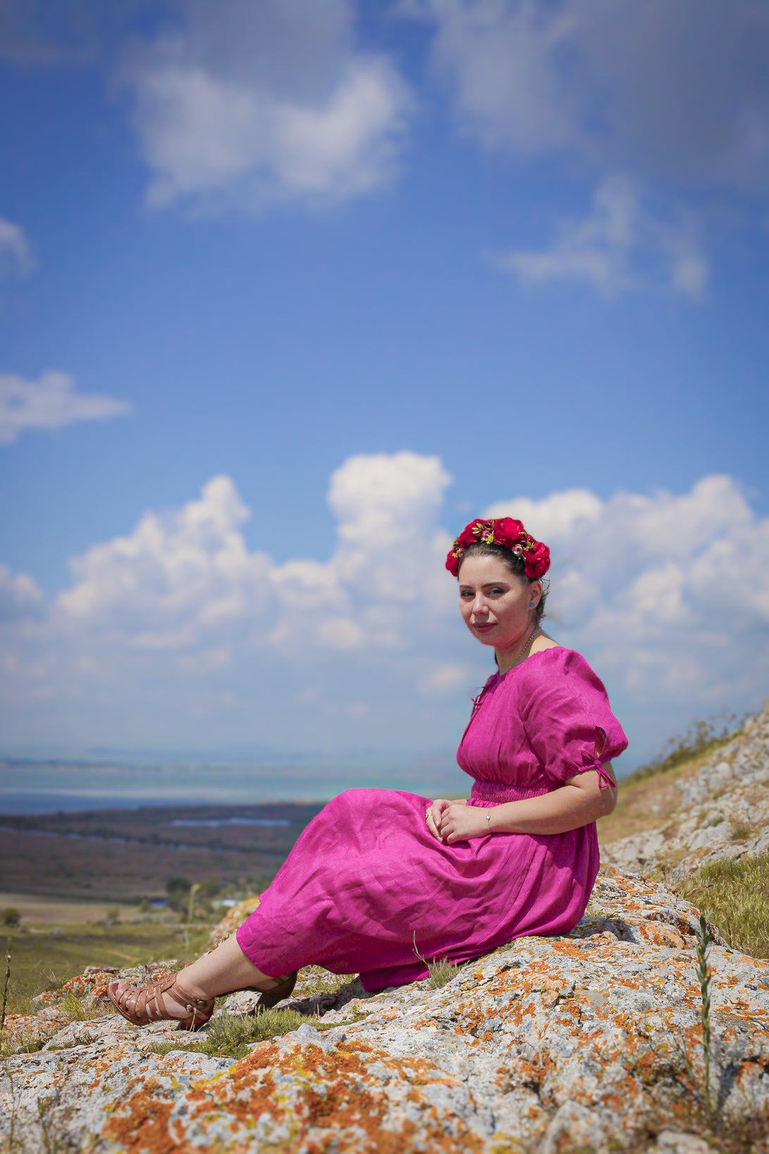 Woman in a pink modest nursing dress sitting on a rocky outcrop with a blue sky and clouds in the background