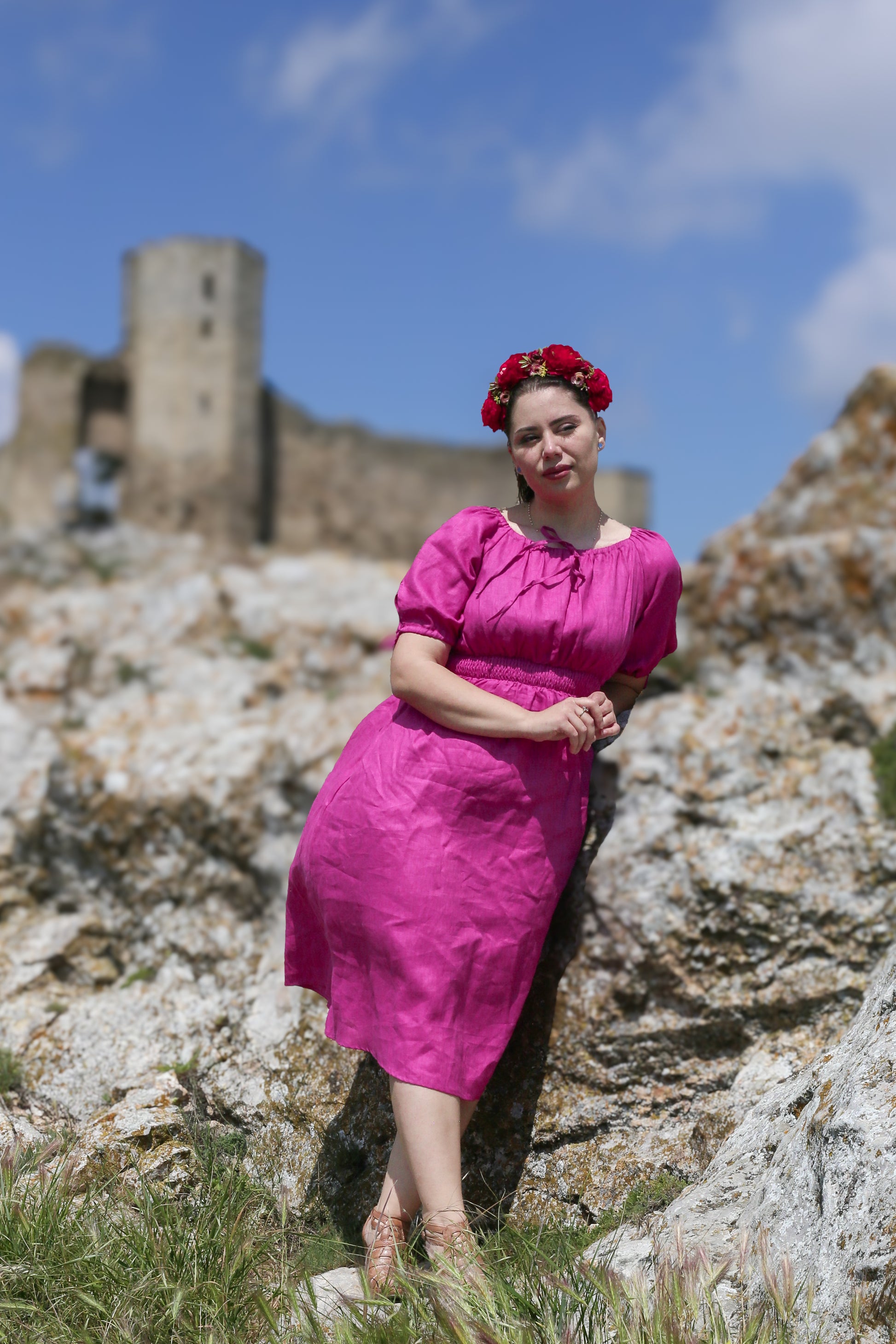 Woman in a pink modest nursing dress standing in front of a castle with rocky terrain and blue sky.
